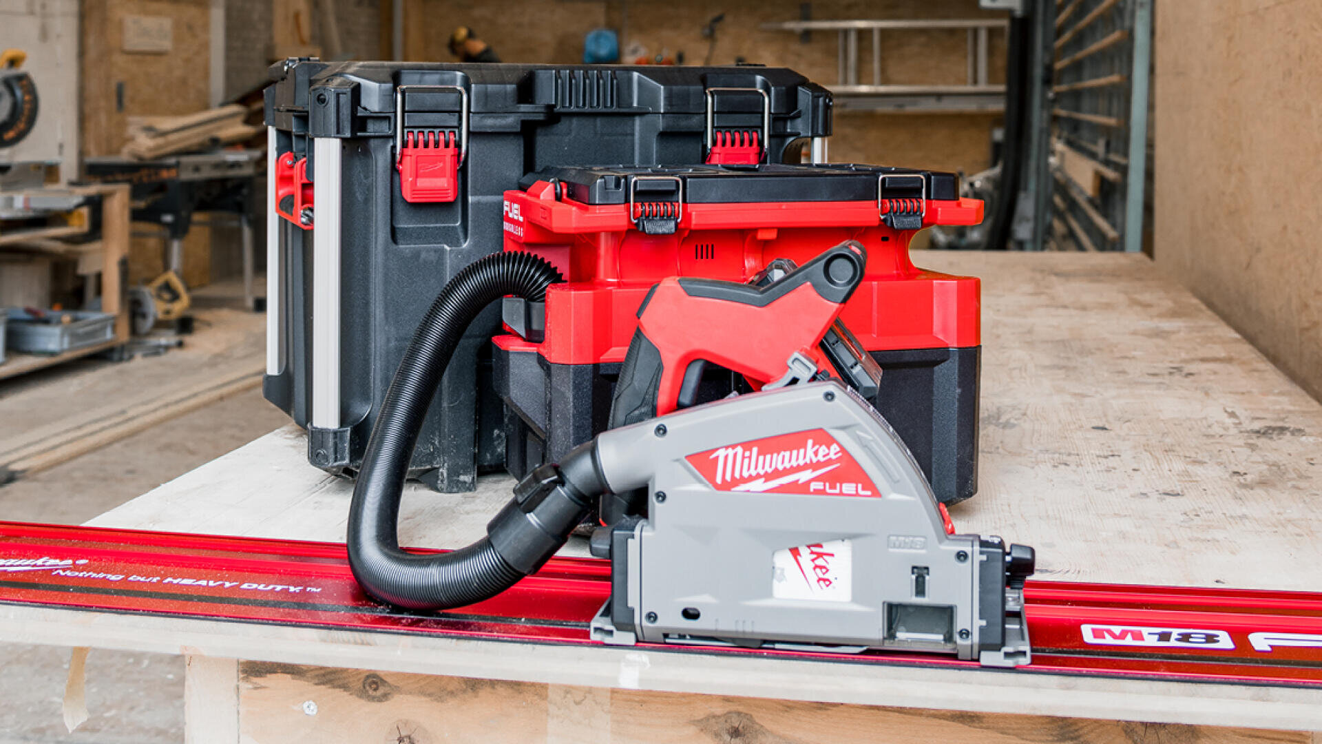 Eine Milwaukee Fuel Kettensäge mit Absaugschlauch ist auf einer roten Führungsschiene positioniert, vor zwei Kisten mit Milwaukee-Werkzeugen, die auf einer Werkbank in einer Werkstatt gestapelt sind.