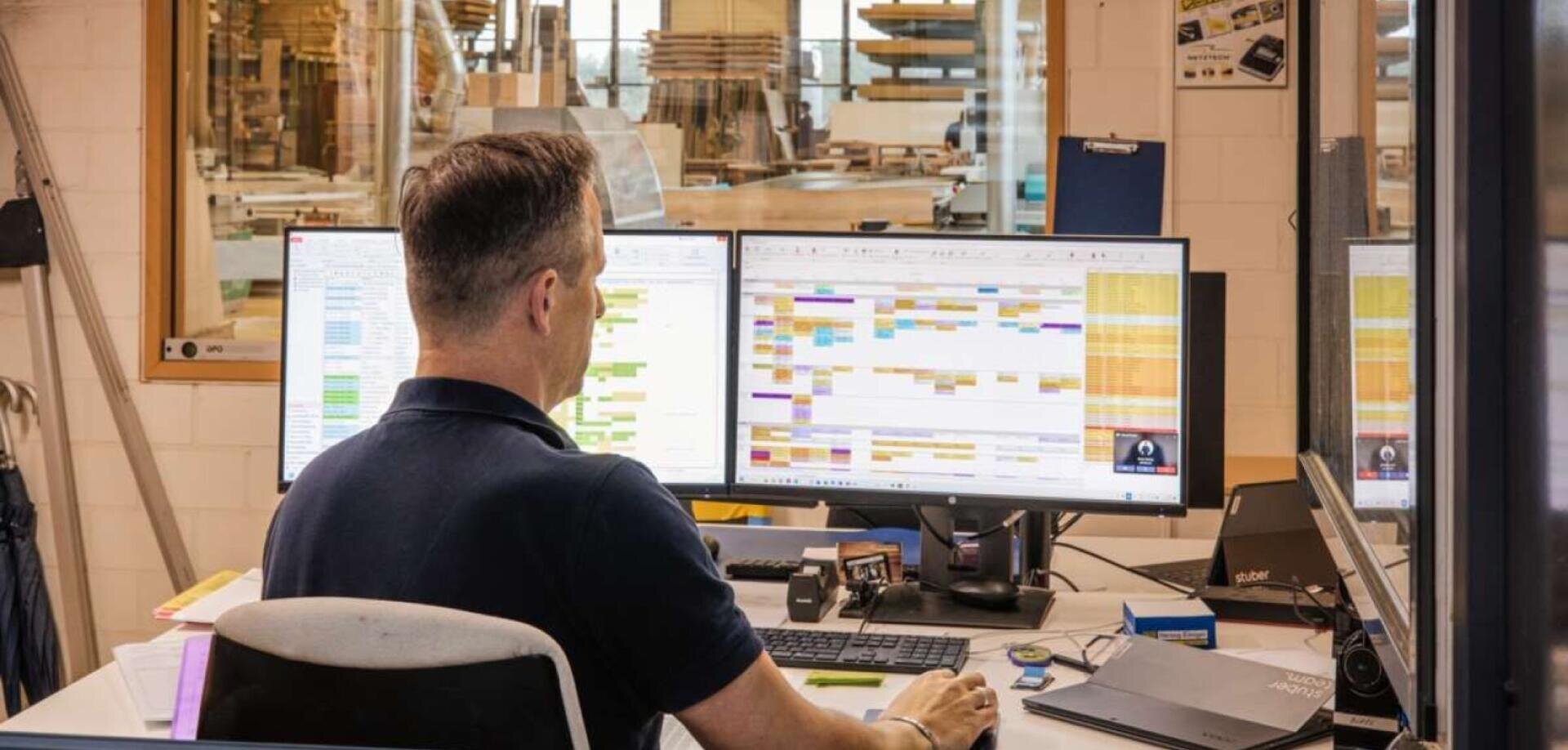 Ein Mann sitzt an einem Schreibtisch in einem Büro und arbeitet an zwei großen Computermonitoren, auf denen bunte Zeitpläne und Tabellenkalkulationen angezeigt werden, wobei der Arbeitsbereich durch ein Fenster hinter ihm sichtbar ist.