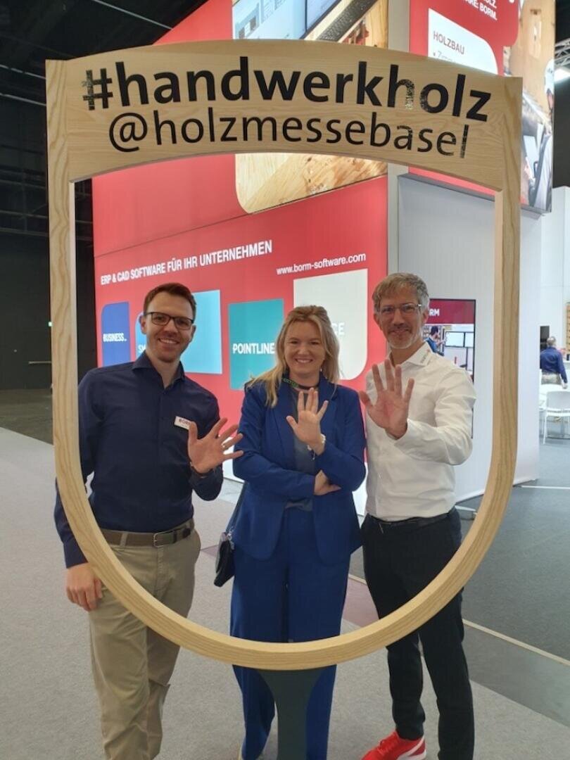 Trois personnes souriantes, les mains en l'air, montrant chacune cinq doigts, alors qu'elles posent derrière un grand cadre en bois portant l'inscription #handwerkholz @holzmessebasel lors d'une exposition ou d'un salon en intérieur.