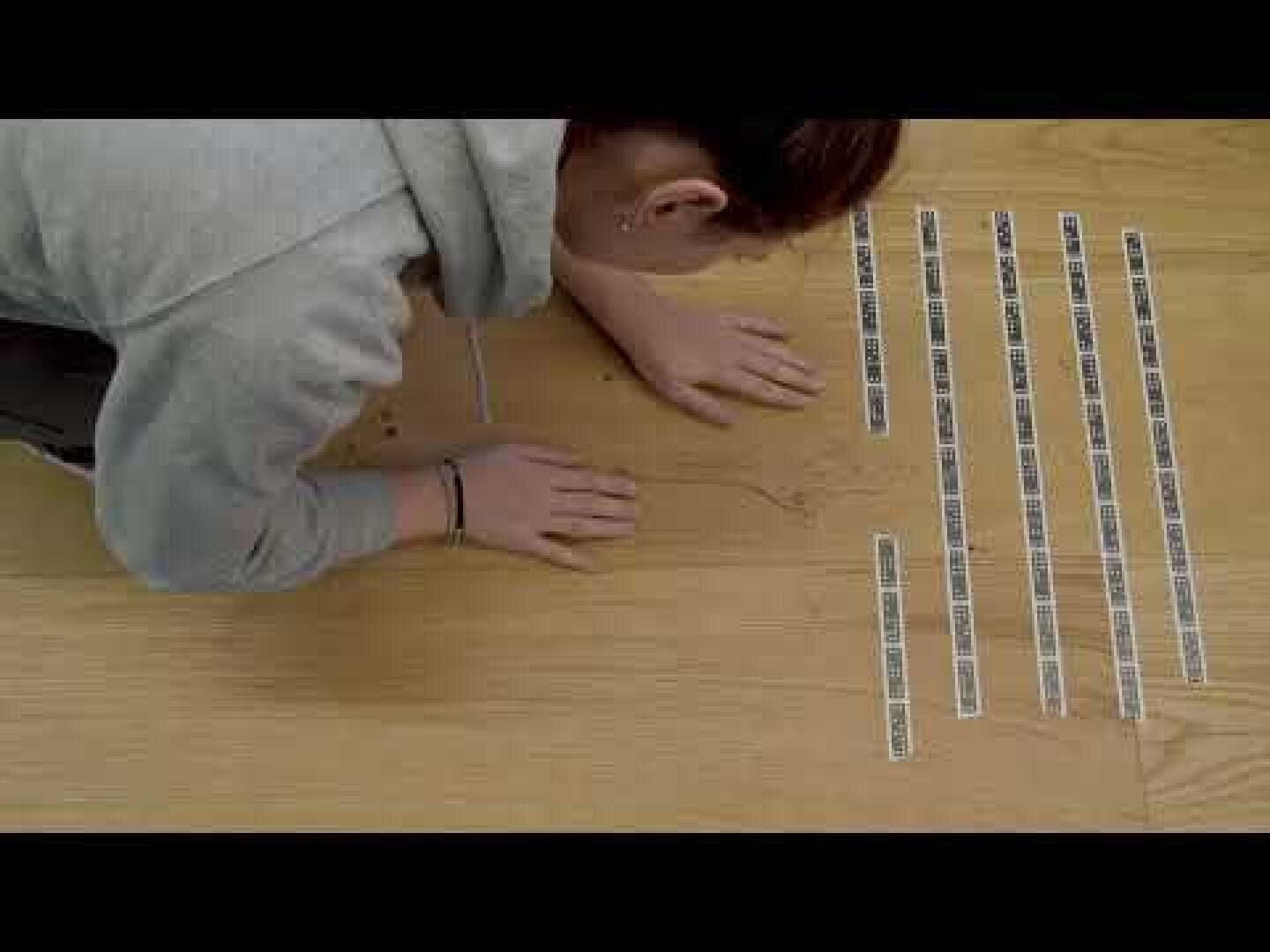 Une personne vêtue d'un sweat à capuche gris est agenouillée sur un sol en bois et examine attentivement une ligne de faible intensité. À proximité, plusieurs bandes portant des codes-barres et du texte sont disposées parallèlement les unes aux autres.