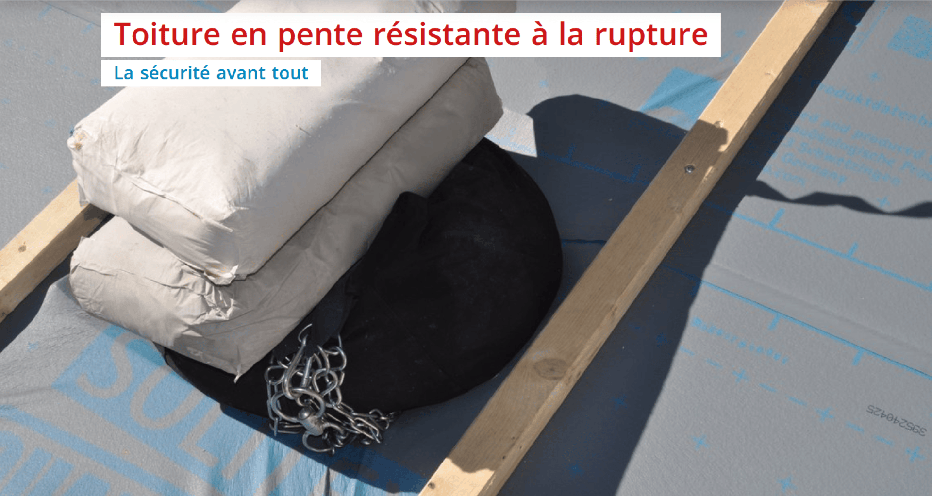 Sacs lestés et chaînes placés sur un toit en pente en cours de construction, avec des poutres en bois et des matériaux de couverture visibles. Le texte en français se lit comme suit : Toiture en pente résistante à la rupture. La sécurité avant tout.