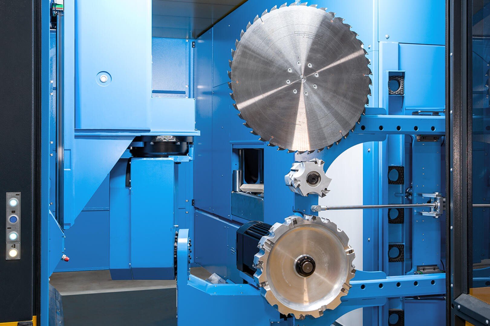 Große Kreissägeblätter, montiert auf einer blauen Industriemaschine aus Metall, mit sichtbaren Hebeln, Schalttafeln und Bauteilen, die auf eine hochpräzise Schneid- oder Fertigungsumgebung hindeuten.