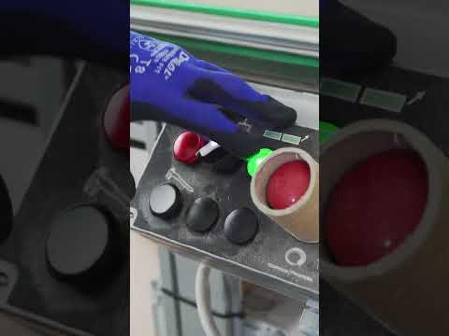 Une personne portant un gant bleu appuie sur un bouton vert lumineux sur un panneau de commande avec plusieurs boutons et cadrans à côté d'un tube en carton contenant un liquide ou une substance rouge.