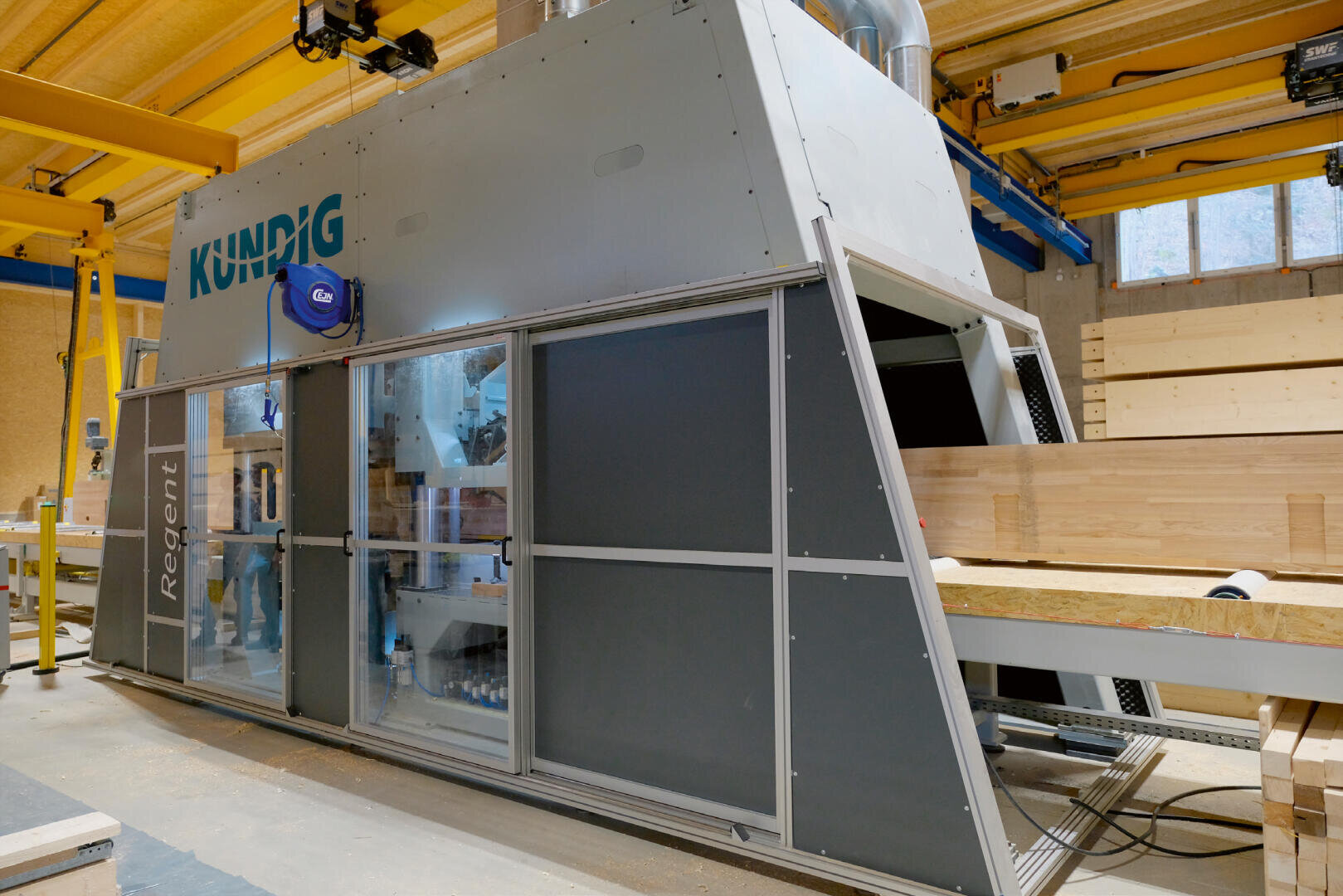 Eine grosse Industrie-Schleifmaschine der Marke Kündig bearbeitet Holzbalken in einer hellen, modernen Holzwerkstatt mit Holzwänden und -decken.