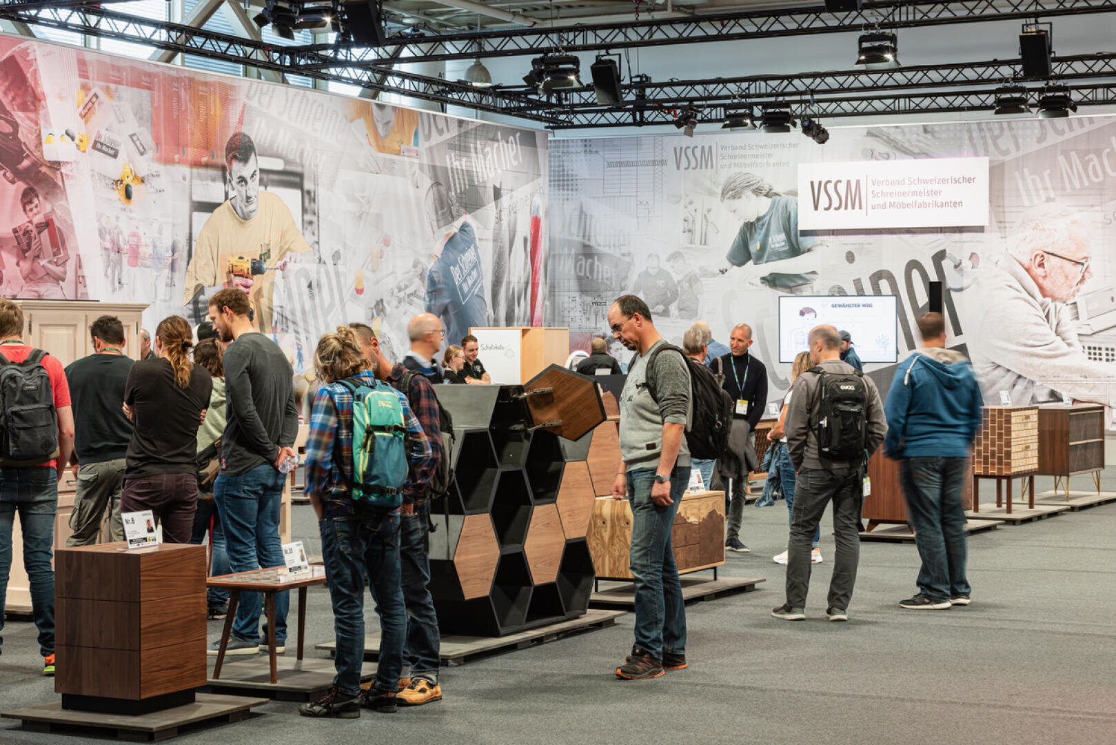 Eine Gruppe von Personen besucht einen Stand auf einer Fachmesse, auf dem Möbel und Displays aus Holz präsentiert werden. Der Stand hat Informationstafeln und ein großes Wandgemälde mit Bildern von Holzarbeiten an der Rückwand. Einige Teilnehmer interagieren mit den Ausstellungsstücken.