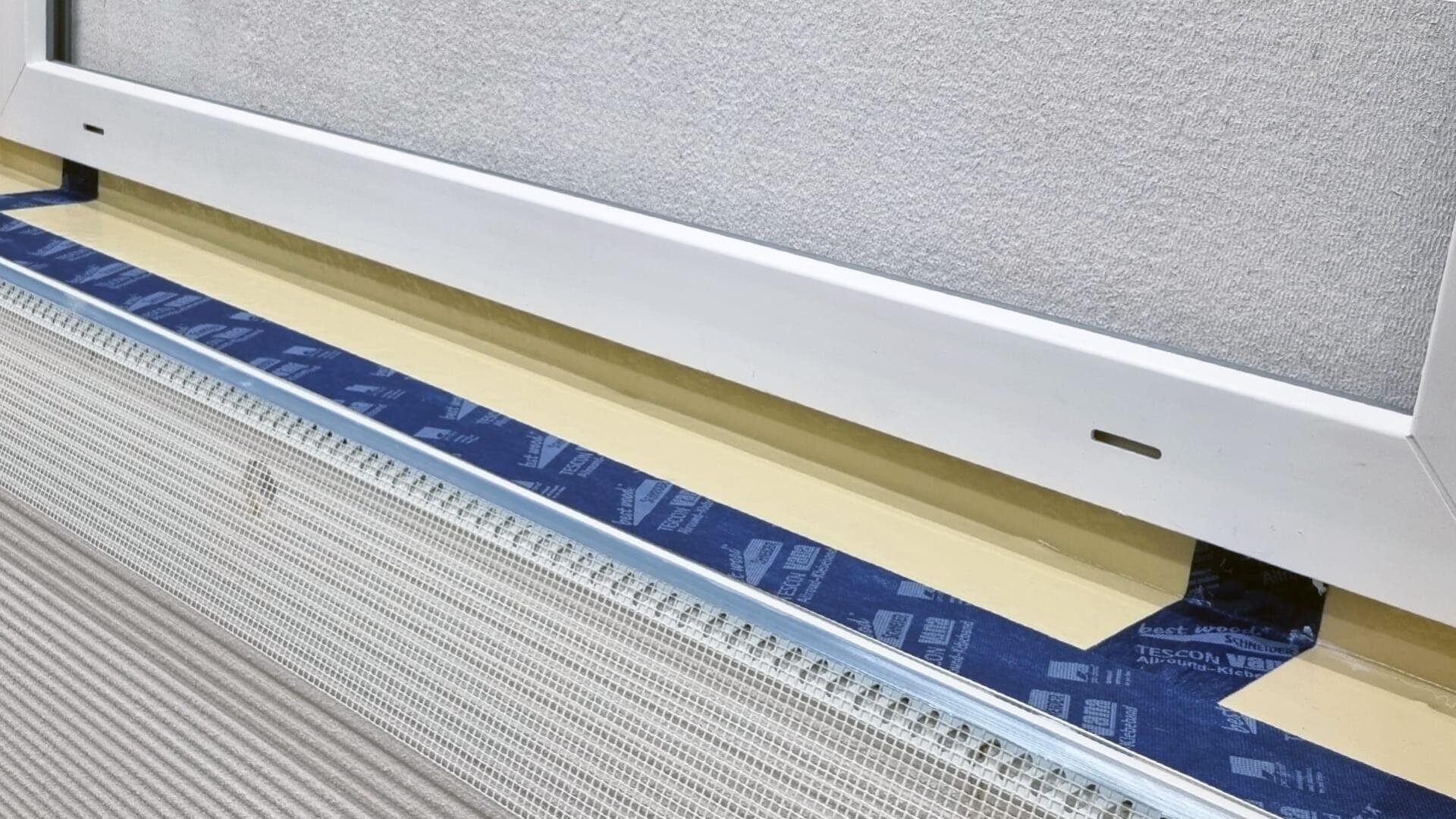 Eine Nahaufnahme einer im Bau befindlichen Wand mit freiliegender Isolierung, einem Metallrahmenstreifen, blauem wasserdichtem Klebeband mit der Aufschrift „TESCON“ und einem weißen Fenster- oder Plattenrahmen darüber.