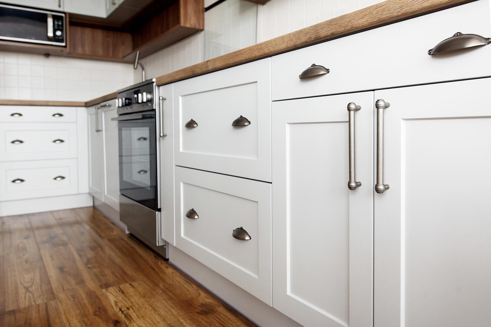 Eine moderne Küche mit weißen Schränken, Griffen aus rostfreiem Stahl, Arbeitsplatten aus Holz, einem Herd aus rostfreiem Stahl, einer Mikrowelle und Holzböden. Das Design ist sauber und minimalistisch.