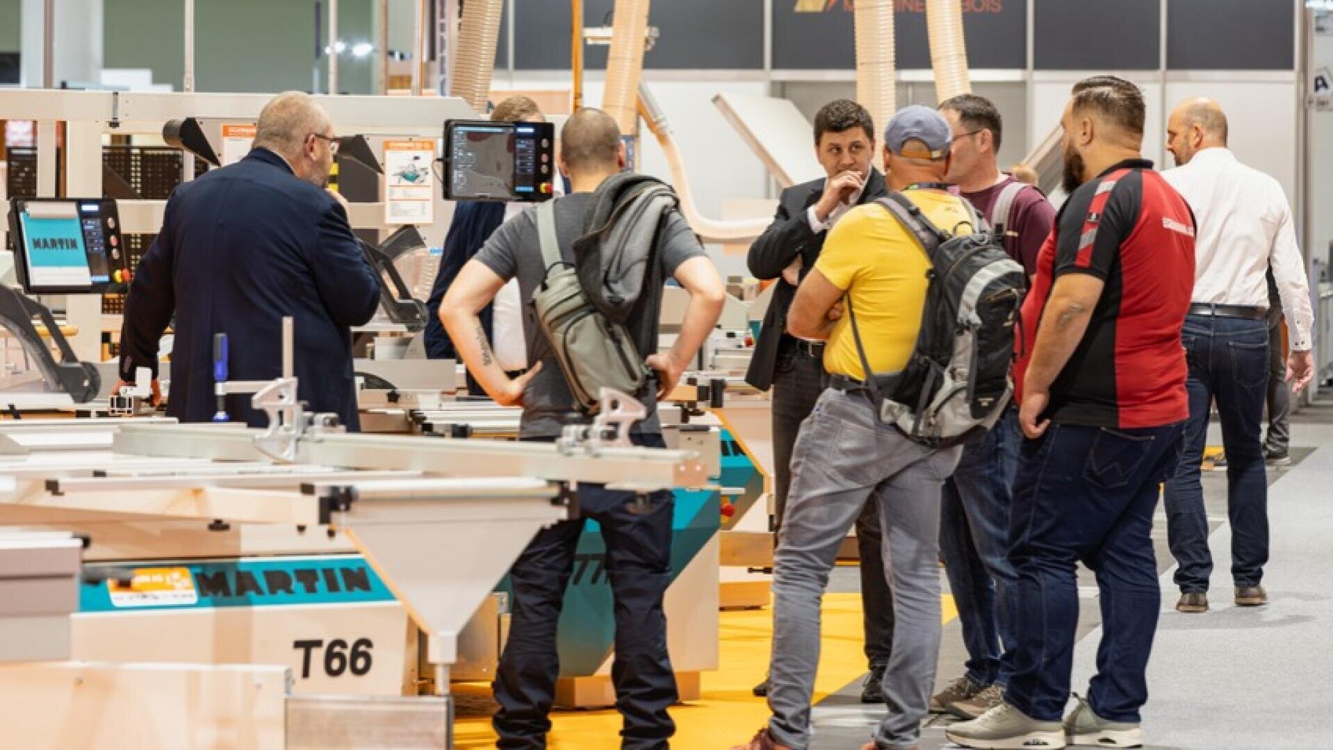 Eine Gruppe von Männern steht auf einer Messe oder Ausstellung um Maschinen herum, unterhält sich und betrachtet die ausgestellten Geräte. Die meisten sind leger gekleidet und tragen Rucksäcke.