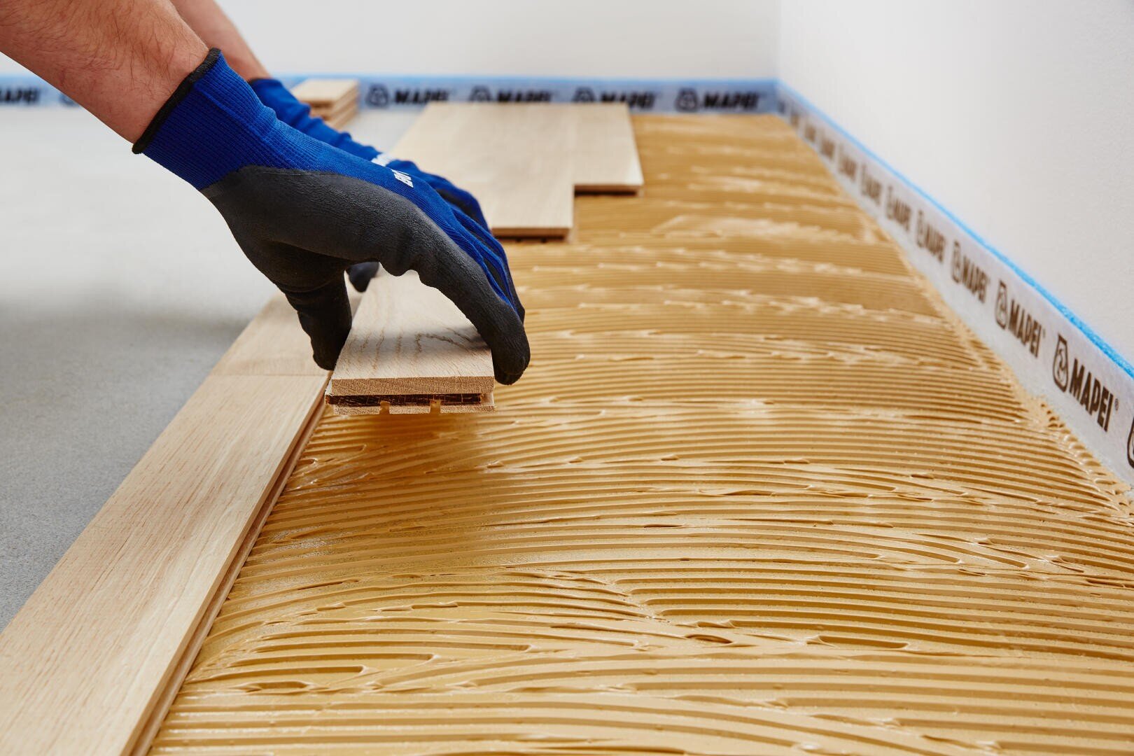 Eine Person mit blauen Handschuhen verlegt Holzdielen über einer Kleberschicht, die gleichmäßig auf dem Boden verteilt wurde.