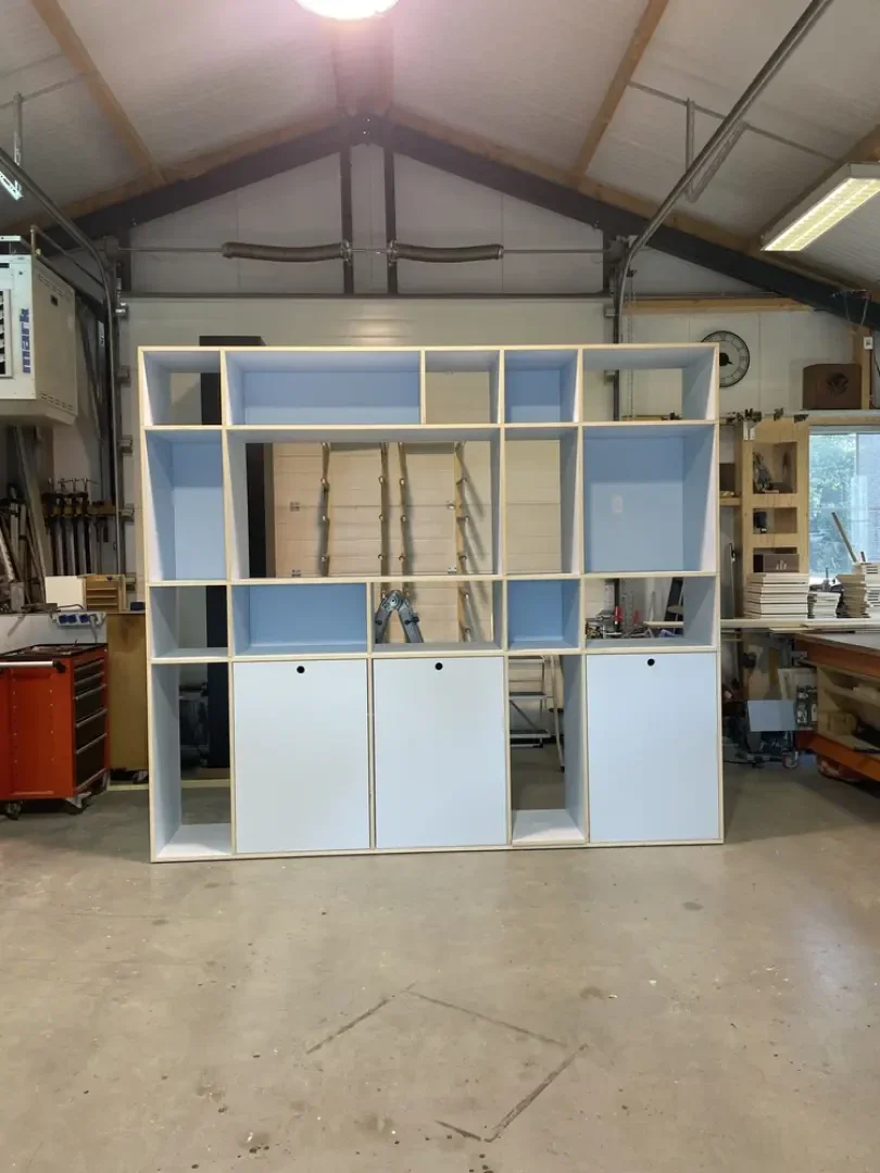 In der Mitte einer Werkstatt steht ein großes, hellblau-weißes Regal mit verschiedenen Fächern und drei unteren Schränken. Im Hintergrund sind Werkzeuge, Geräte und Fenster zu sehen.