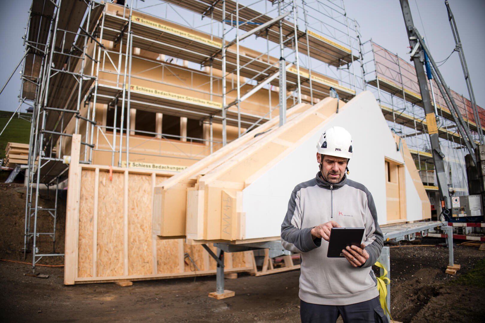 Ein Bauarbeiter in einer grauen Jacke und einem weißen Schutzhelm benutzt ein Tablet auf einer Baustelle mit Gerüsten, Holzstrukturen und Baumaterialien im Hintergrund.