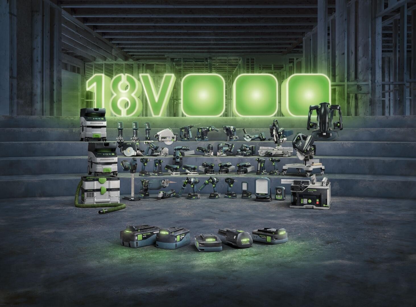 Ein Display mit verschiedenen 18-V-Elektrowerkzeugen und Zubehör von Festool, angeordnet auf Stufen in einem teilweise errichteten Gebäude, mit einem leuchtenden grünen 18-V-Schild dahinter und Batterien im Vordergrund.