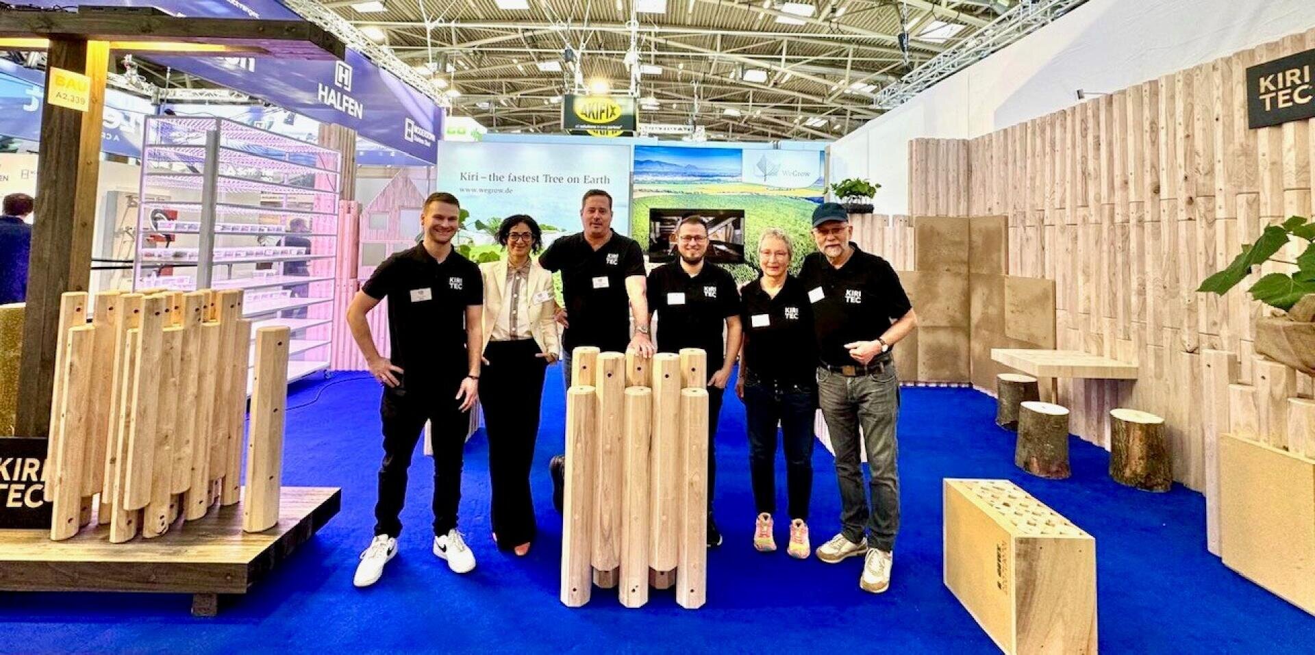 Sechs Personen stehen lächelnd an einem Messestand mit hölzernen Ausstellungsstücken, blauem Teppich und Werbebannern im Hintergrund. Die Gruppe trägt zusammenpassende schwarze Hemden, mit Ausnahme einer Person in Weiß, und der Stand hat ein naturbezogenes Design.