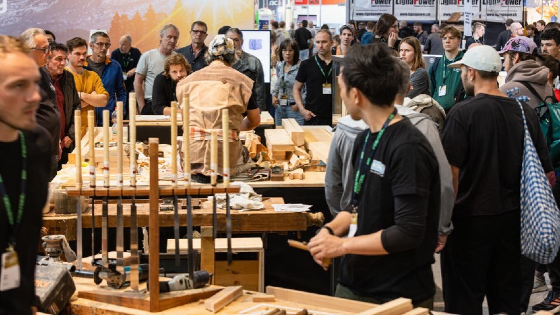 Une foule assiste à des démonstrations de travail du bois lors d'un événement très fréquenté. Les gens se tiennent autour d'établis avec des outils, des pièces de bois et des serre-joints, tandis qu'une personne en tablier travaille au centre. L'atmosphère est animée et concentrée.