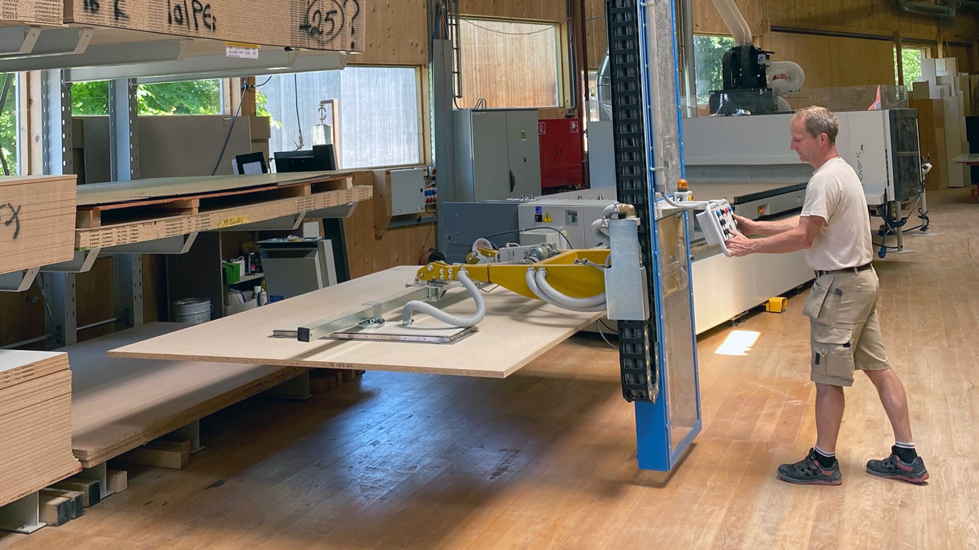 Eine Person bedient eine große blaue Industriemaschine, die eine Platte in einer Holzbearbeitungswerkstatt anhebt, wobei gestapelte Holzplatten und Geräte sichtbar sind.
