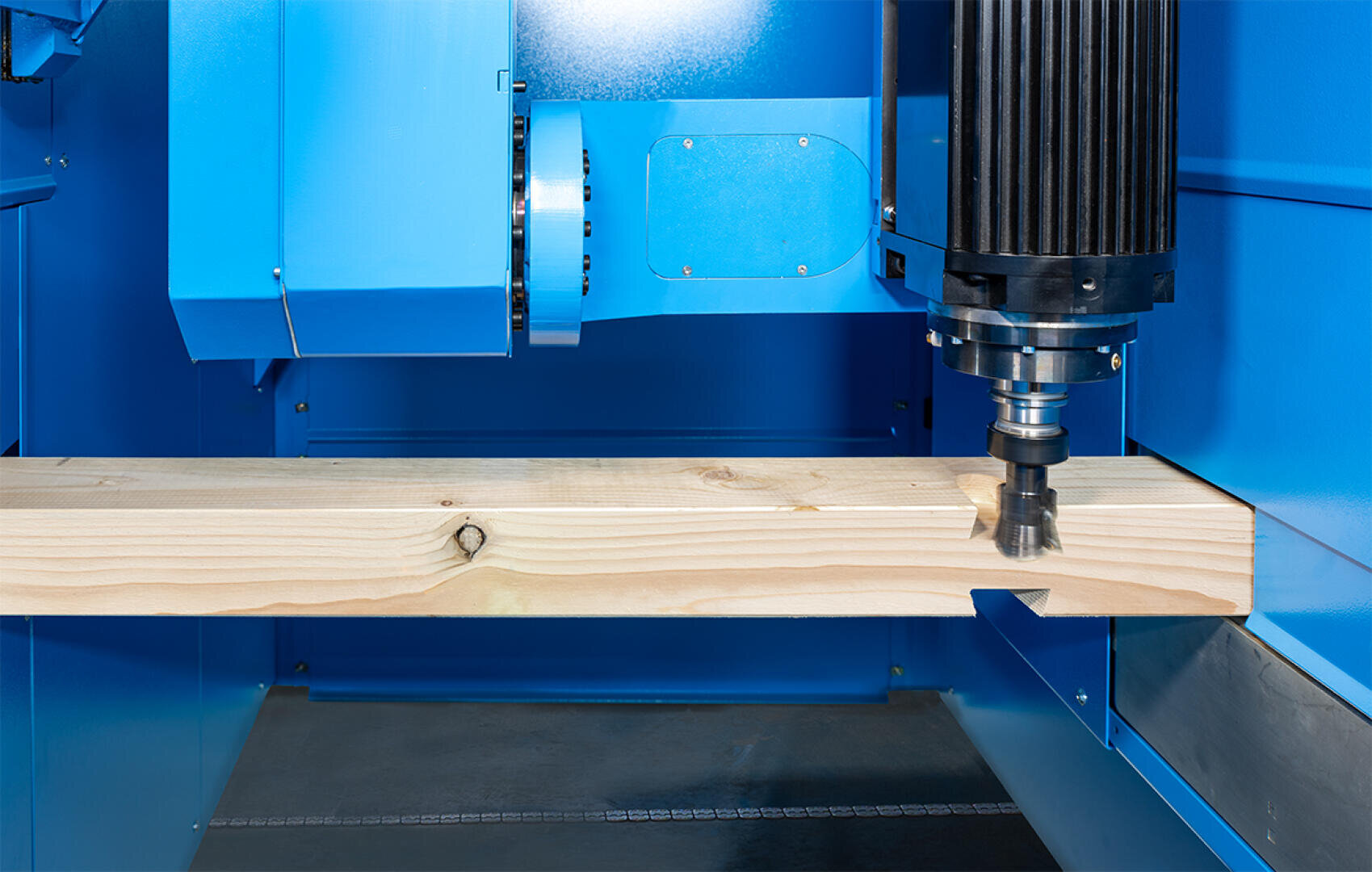 Nahaufnahme einer CNC-Fräsmaschine, die ein Stück helles Holz fräst, wobei die Maschinenteile blau lackiert sind und das Fräswerkzeug das Holz berührt.