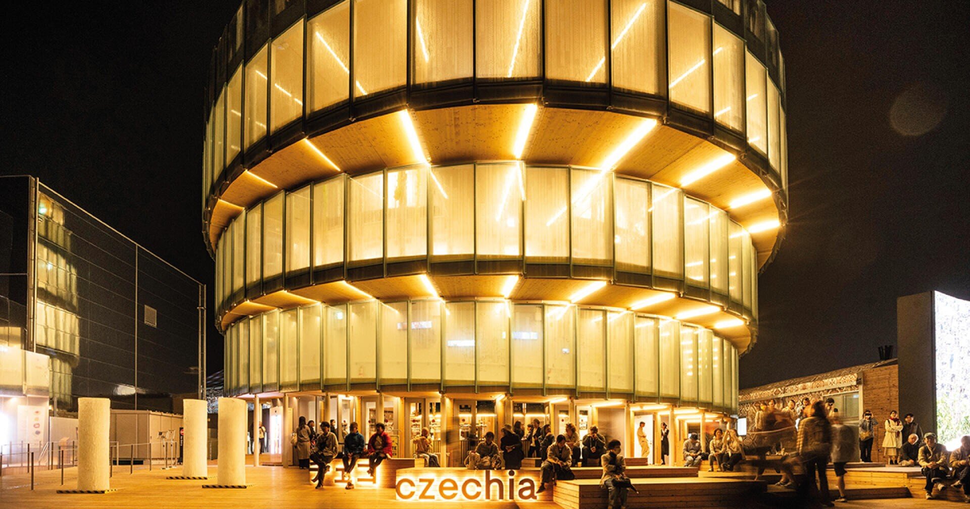 Ein modernes, rundes Glasgebäude mit beleuchteten Tafeln bei Nacht. Menschen sind um den Eingang versammelt, und das Wort czechia leuchtet in orangefarbenen Buchstaben am Sockel.