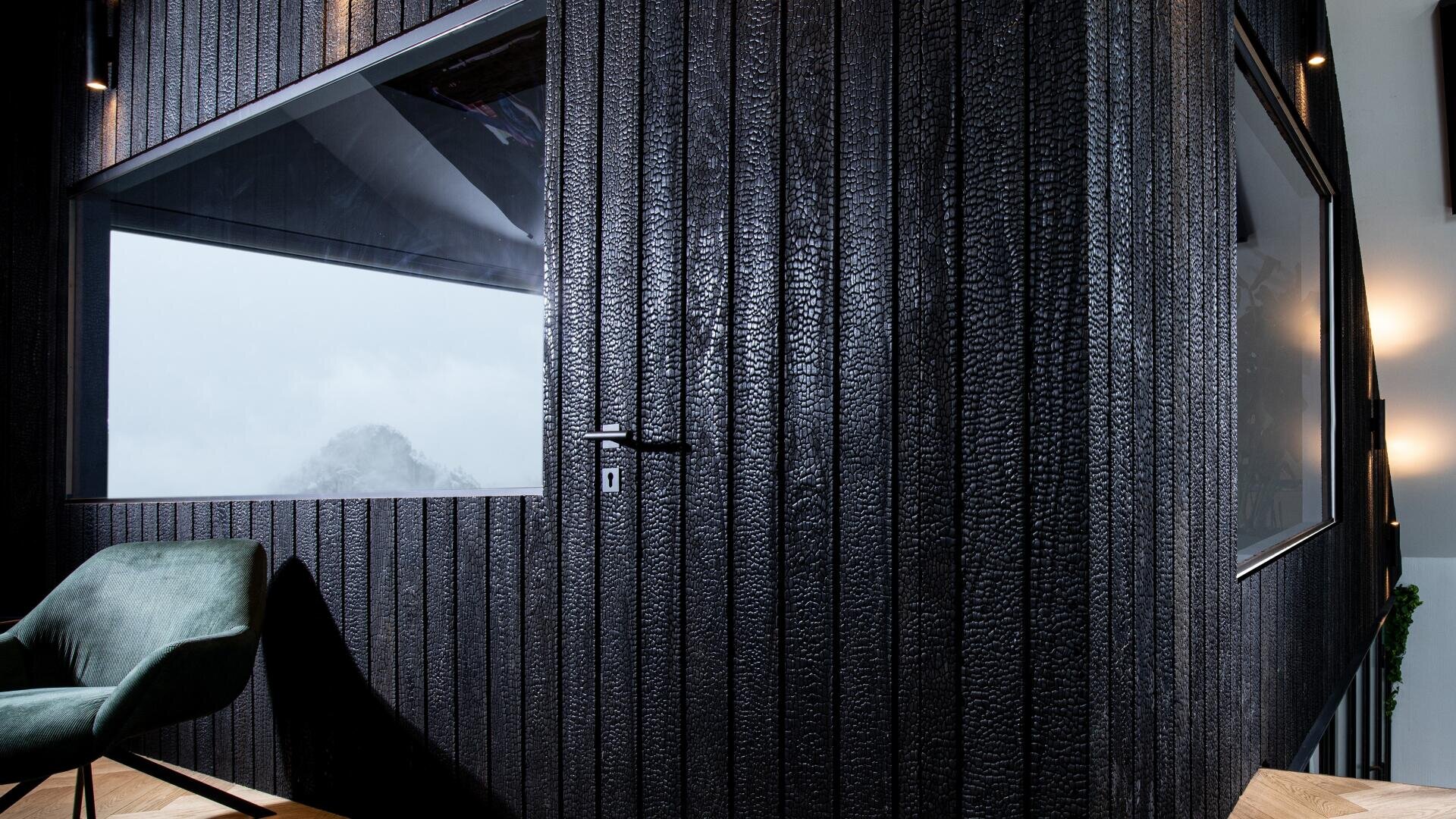 Eine moderne Zimmerecke mit schwarzen, strukturierten Holzwänden und großen Fenstern. Ein grün gepolsterter Stuhl steht in der Nähe, und durch das Fenster sind neblige Berge zu sehen. Der Fußboden hat ein helles Holz-Fischgrätenmuster.