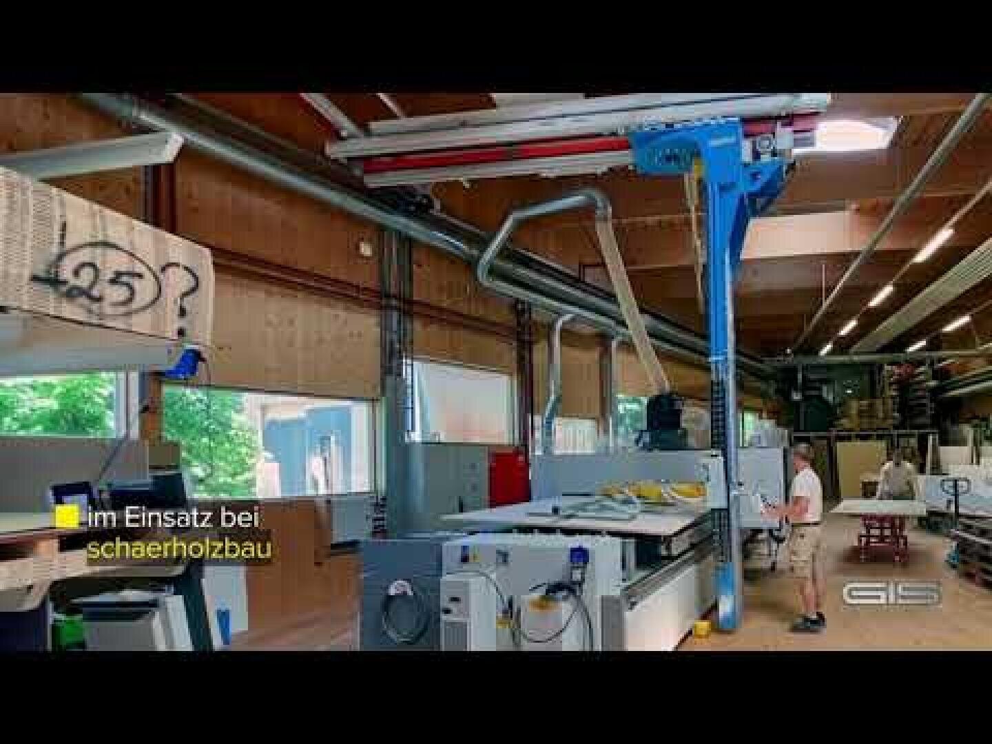 Un atelier de menuiserie spacieux avec différentes machines et appareils. Une personne se tient à proximité d'un grand poste de travail. Des poutres en bois et des fenêtres sont visibles. En allemand, on peut lire en jaune : "Im Einsatz bei schaerholzbau.