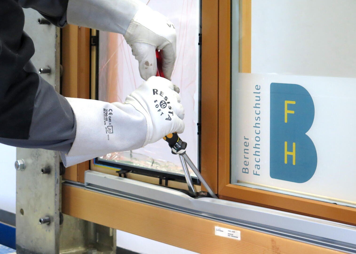 Eine Person mit weißen Handschuhen benutzt eine Zange, um den Rahmen eines Fensters anzupassen. Das Fenster trägt ein blau-gelbes Schild mit der Aufschrift Berner Fachhochschule FH.