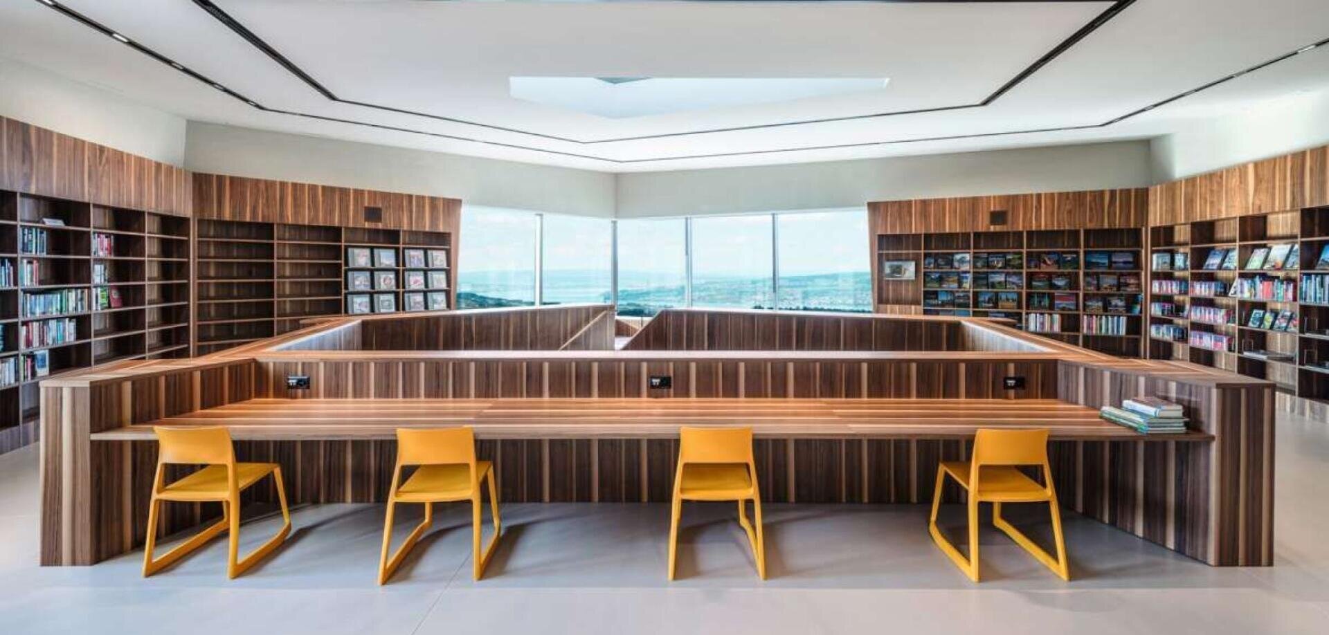 Moderne Bibliothek mit hölzernen Bücherregalen an den Wänden, großen Fenstern mit Blick auf die Landschaft und einem zentralen hölzernen Schreibtisch mit vier gelben Stühlen. Die Bücher sind im gesamten Raum ordentlich angeordnet.