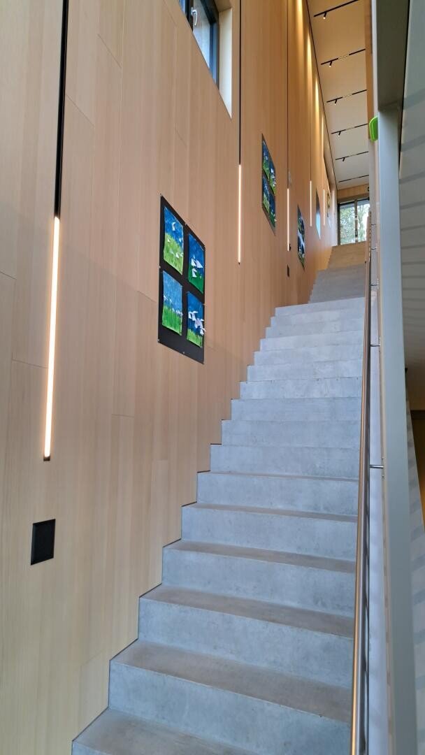 Eine moderne Innentreppe mit elegantem Handlauf führt an einer hohen Holzwand entlang, die mit Kinderzeichnungen dekoriert ist. Vertikale Leuchten sind an der Wand angebracht, und hohe Fenster sorgen für natürliches Licht.