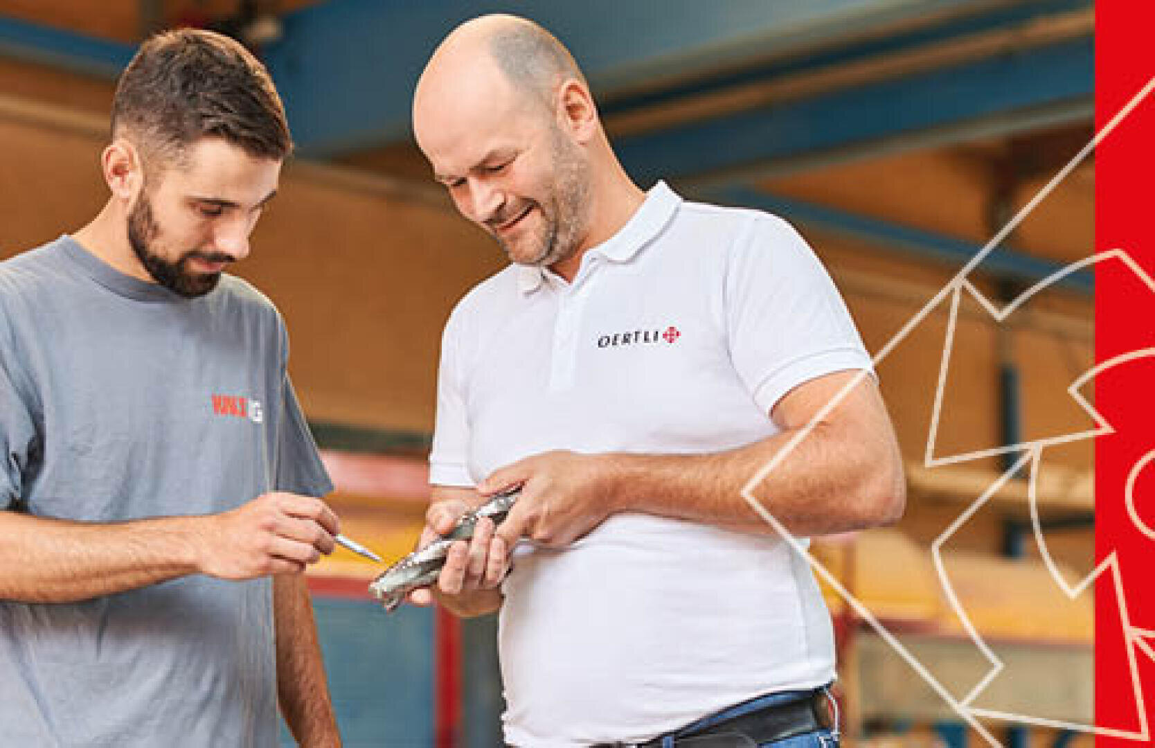 Zwei Männer stehen darin und untersuchen ein mechanisches Teil; ein Mann in einem grauen Hemd zeigt mit einem Bleistift darauf, während der andere in einem weißen Poloshirt lächelt und das Teil hält. Am rechten Rand sind rote geometrische Muster zu sehen.