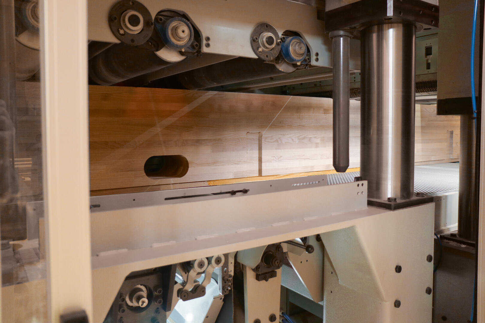 Nahaufnahme einer Industriemaschine, die ein Holzbrett bearbeitet, mit Rollen, Metallteilen und einem teilweise sichtbaren Förderband.