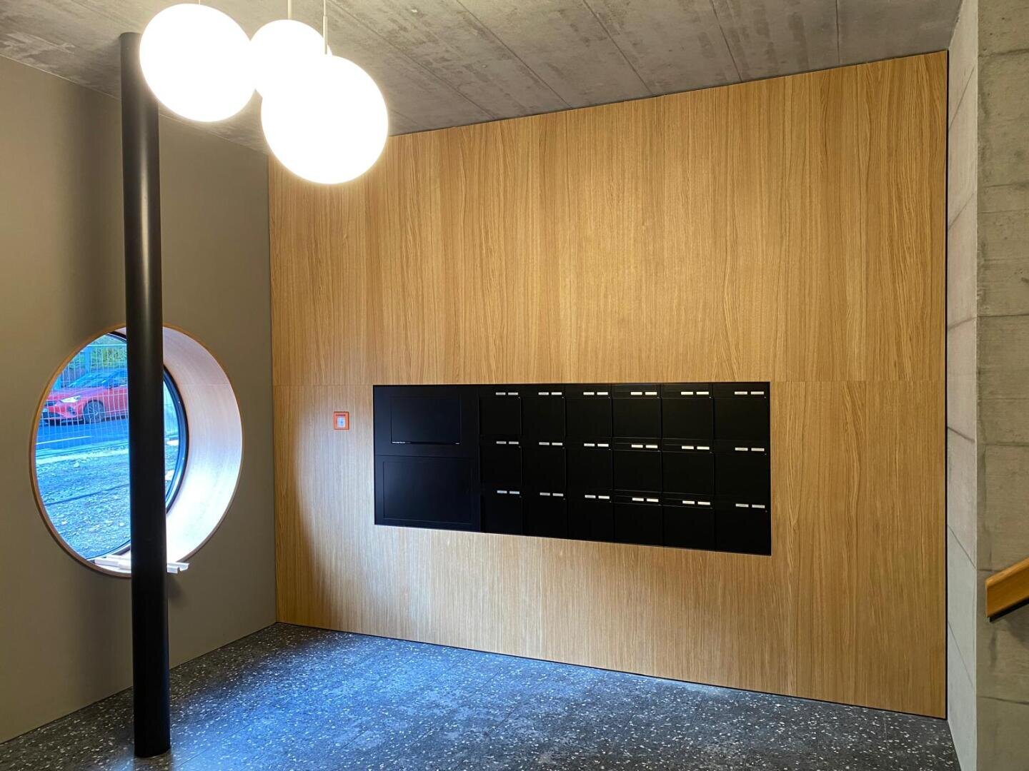 Ein moderner Postraum mit einer holzgetäfelten Wand, schwarzen Briefkästen, einem runden Fenster auf der linken Seite, drei leuchtenden kugelförmigen Deckenleuchten und einem gesprenkelten dunklen Boden.