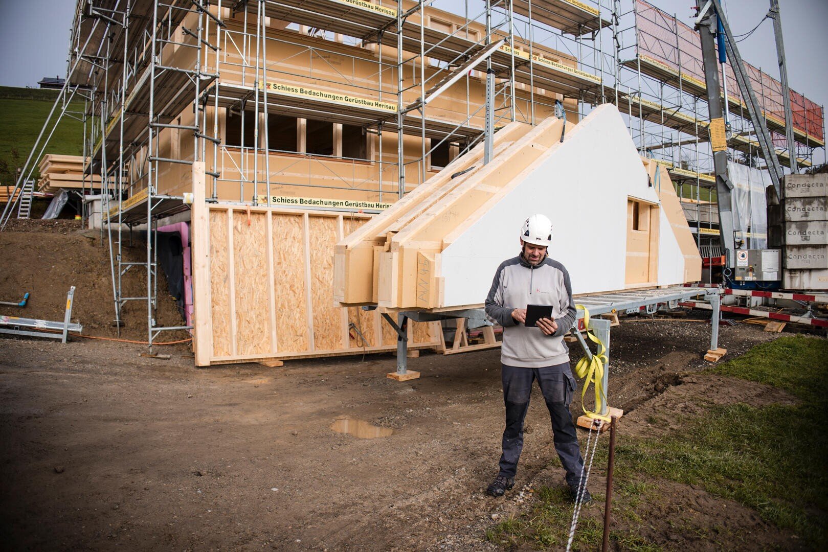 Un ouvrier du bâtiment portant un casque blanc et une veste grise utilise une tablette sur un chantier, avec des échafaudages et une grande structure préfabriquée en bois à l'arrière-plan.