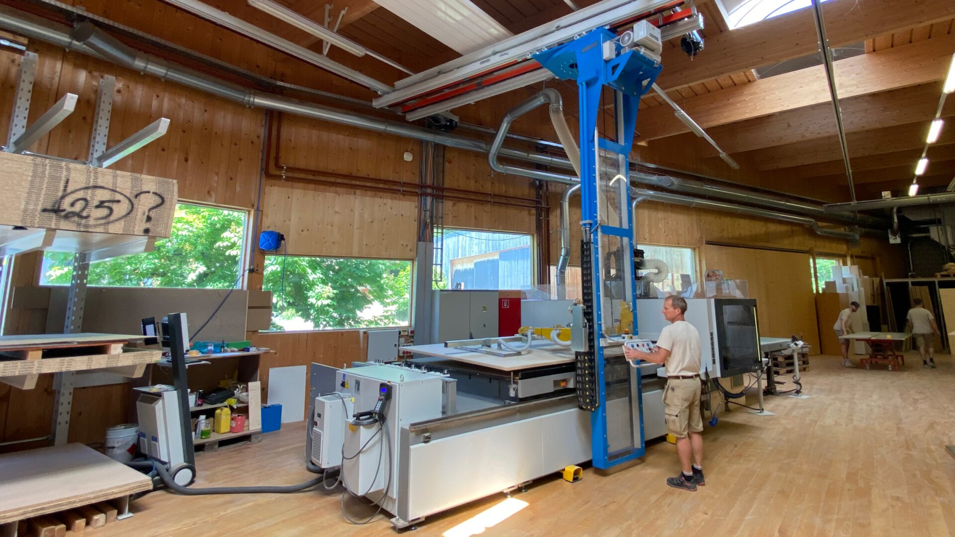 Un homme fait fonctionner une grande machine industrielle bleue et grise dans un atelier spacieux, éclairé par le soleil, avec des murs et des planchers en bois. Une autre personne travaille à l'arrière-plan, près d'étagères et de tables.