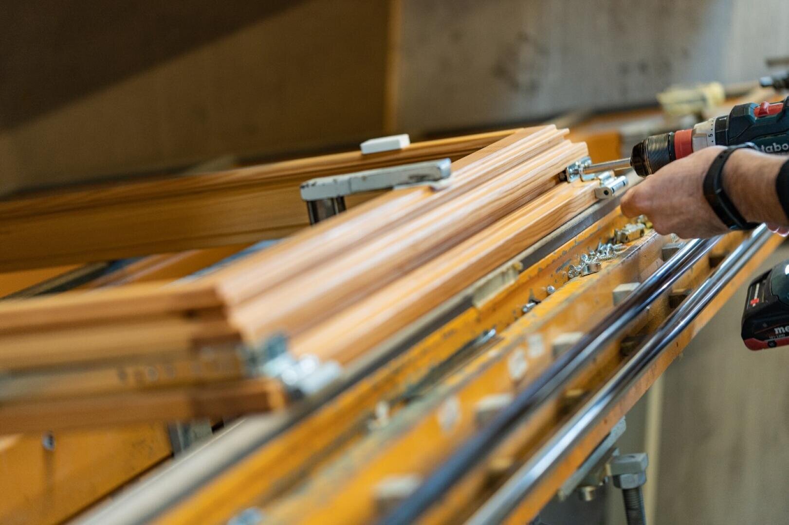 Nahaufnahme einer Person, die eine Bohrmaschine benutzt, um Holzrahmen oder -balken in einer Werkstatt oder in der Fertigung zu montieren oder anzupassen. Auf der Werkbank sind verschiedene Werkzeuge und Metallteile zu sehen.