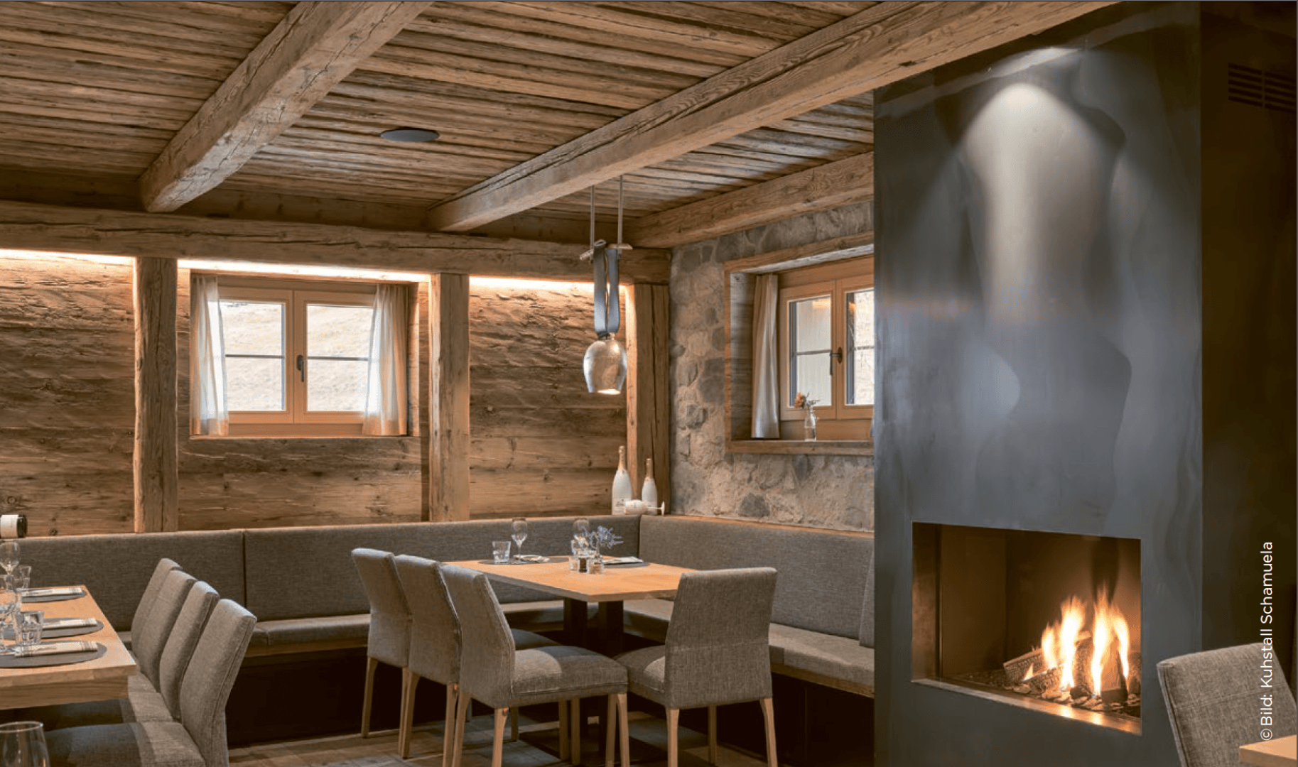 Ein gemütliches Restaurant mit Holzbalken, Steinwänden, modernen grauen Stühlen, gedeckten Tischen und einem beleuchteten Kamin, der eine warme und einladende Atmosphäre schafft. Sonnenlicht strömt durch kleine Fenster.