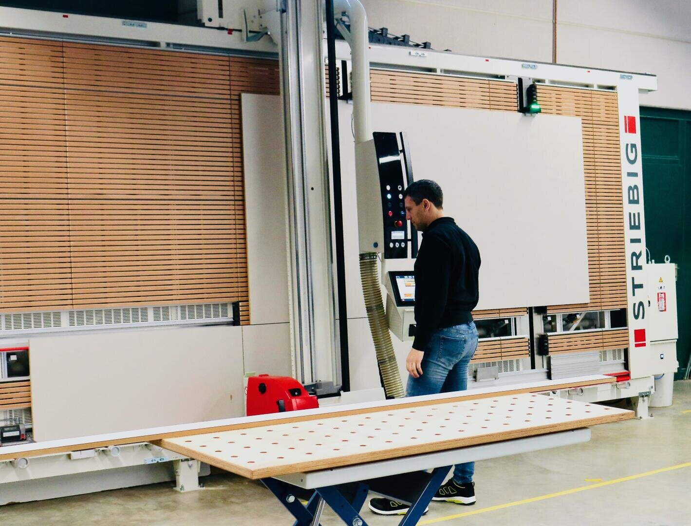 Un homme utilise une grande scie à panneaux verticale dans un atelier et découpe un panneau de bois posé sur une table de travail bleue. La machine porte l'inscription "STRIEBIG".