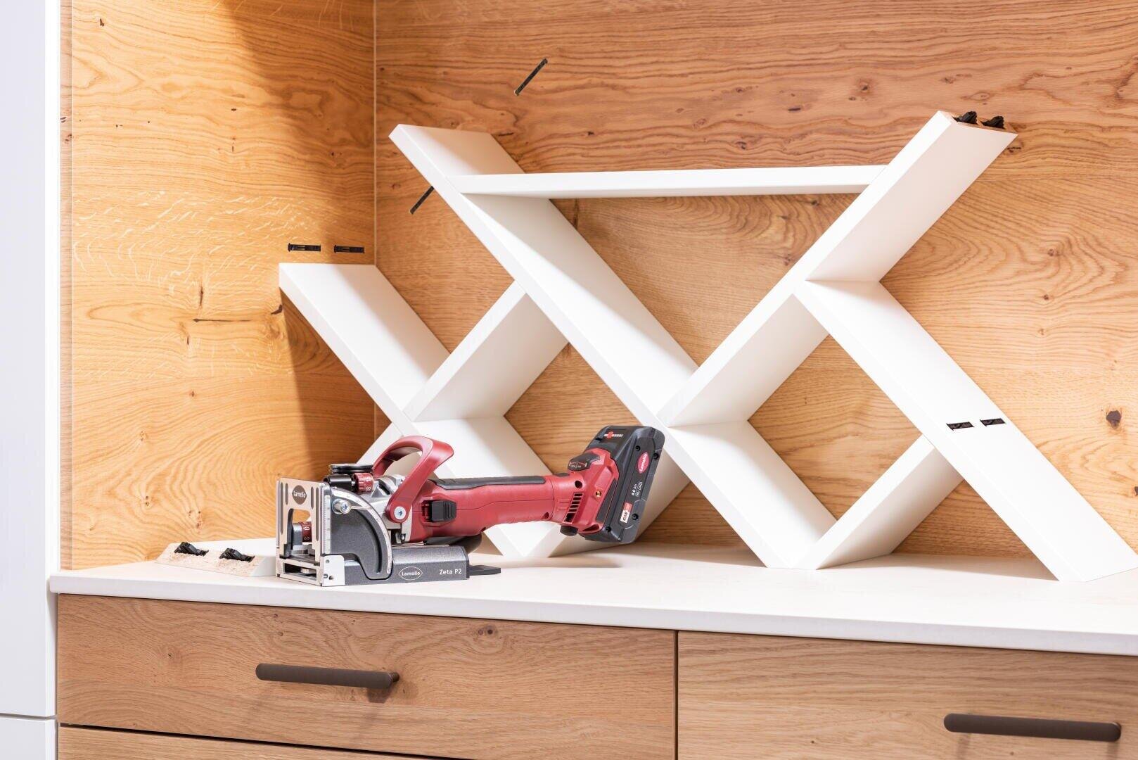 Ein weißes, X-förmiges Bücherregal wird auf einer Arbeitsplatte zusammengebaut, mit einem Tischlerwerkzeug und Schrauben in der Nähe, vor dem Hintergrund einer Holzwand.