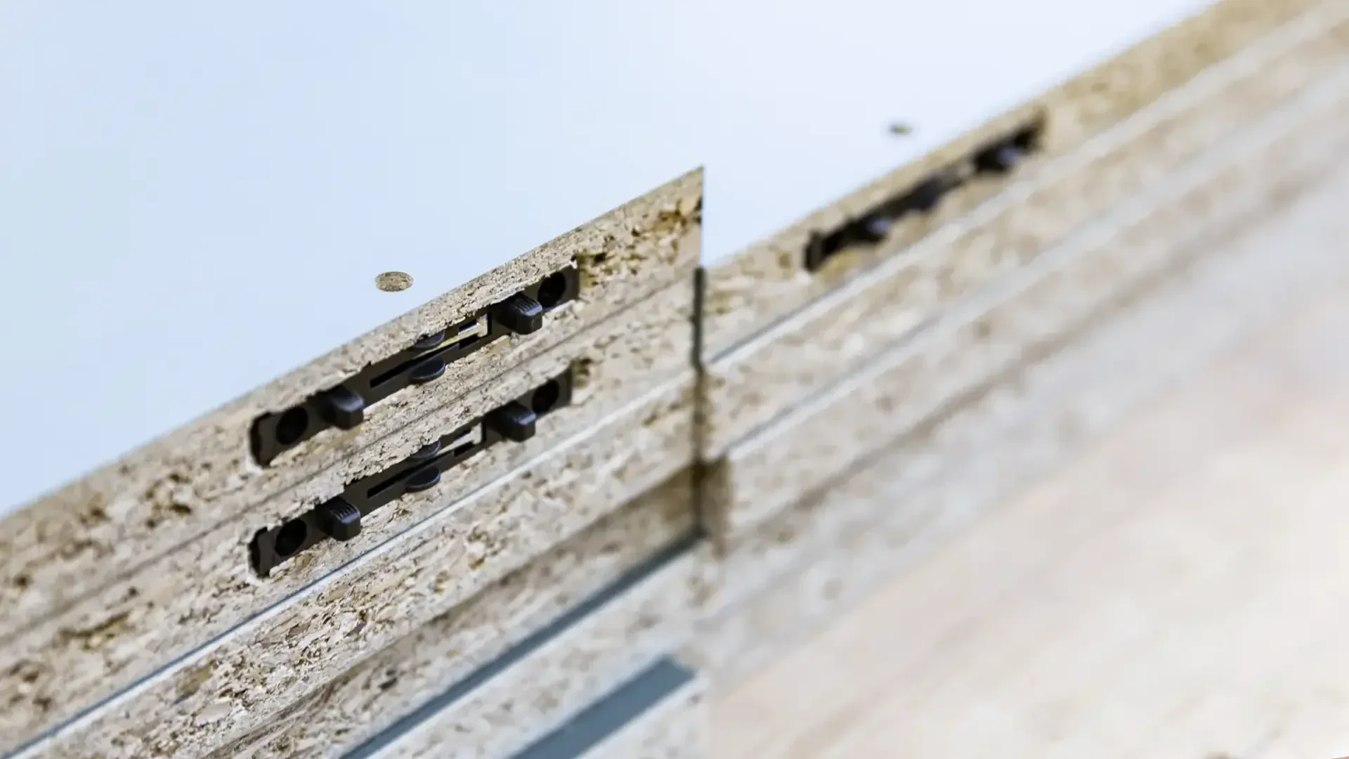 Nahaufnahme der Kante von zwei gestapelten Spanplatten mit verdeckten Metallverbindern, die auf einer Holzoberfläche liegen. Die Paneele sind auf der Oberseite mit einer weißen Beschichtung versehen.