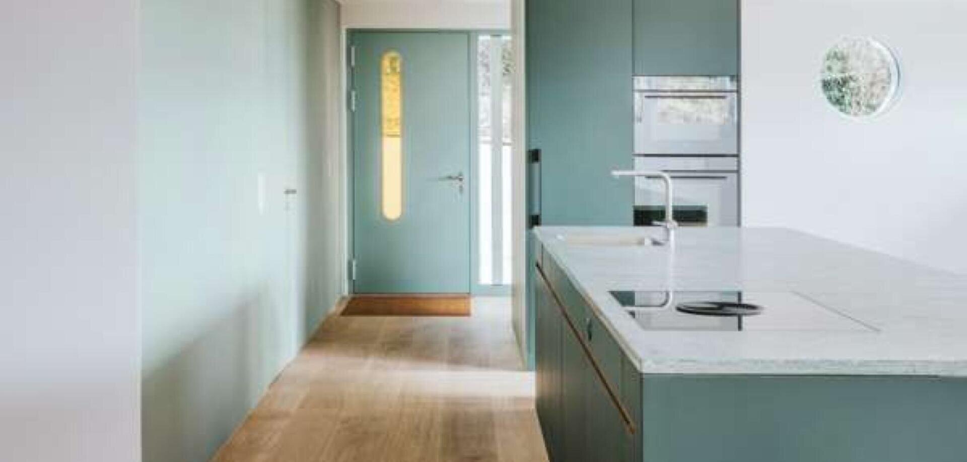Eine moderne Küche mit mintgrünen Schränken, einer Arbeitsplatte mit Marmorinsel, einem eingebauten Ofen und einem Induktionskochfeld. Ein heller Holzfußboden führt zu einer passenden mintgrünen Eingangstür mit einem hohen, schmalen Fenster.