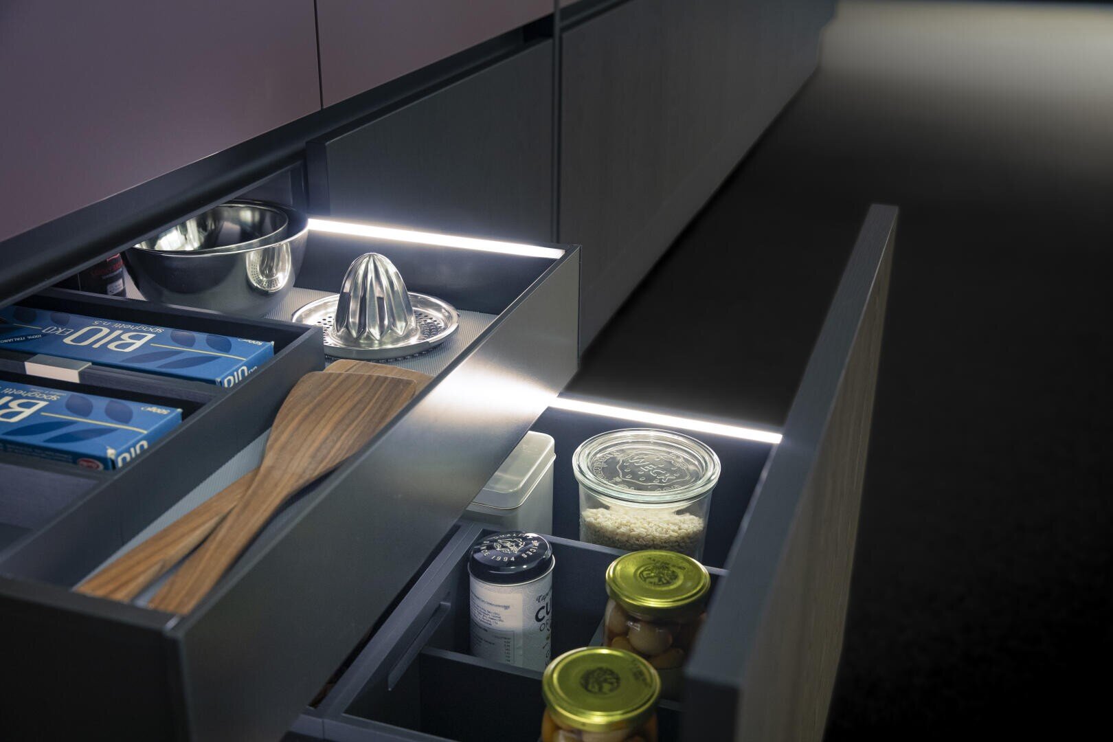 Des tiroirs de cuisine ouverts avec des ustensiles, des bocaux de stockage, un presse-agrumes, des spatules en bois et du papier aluminium à l'intérieur. Des lampes LED éclairent l'intérieur et soulignent l'organisation et l'aménagement moderne.