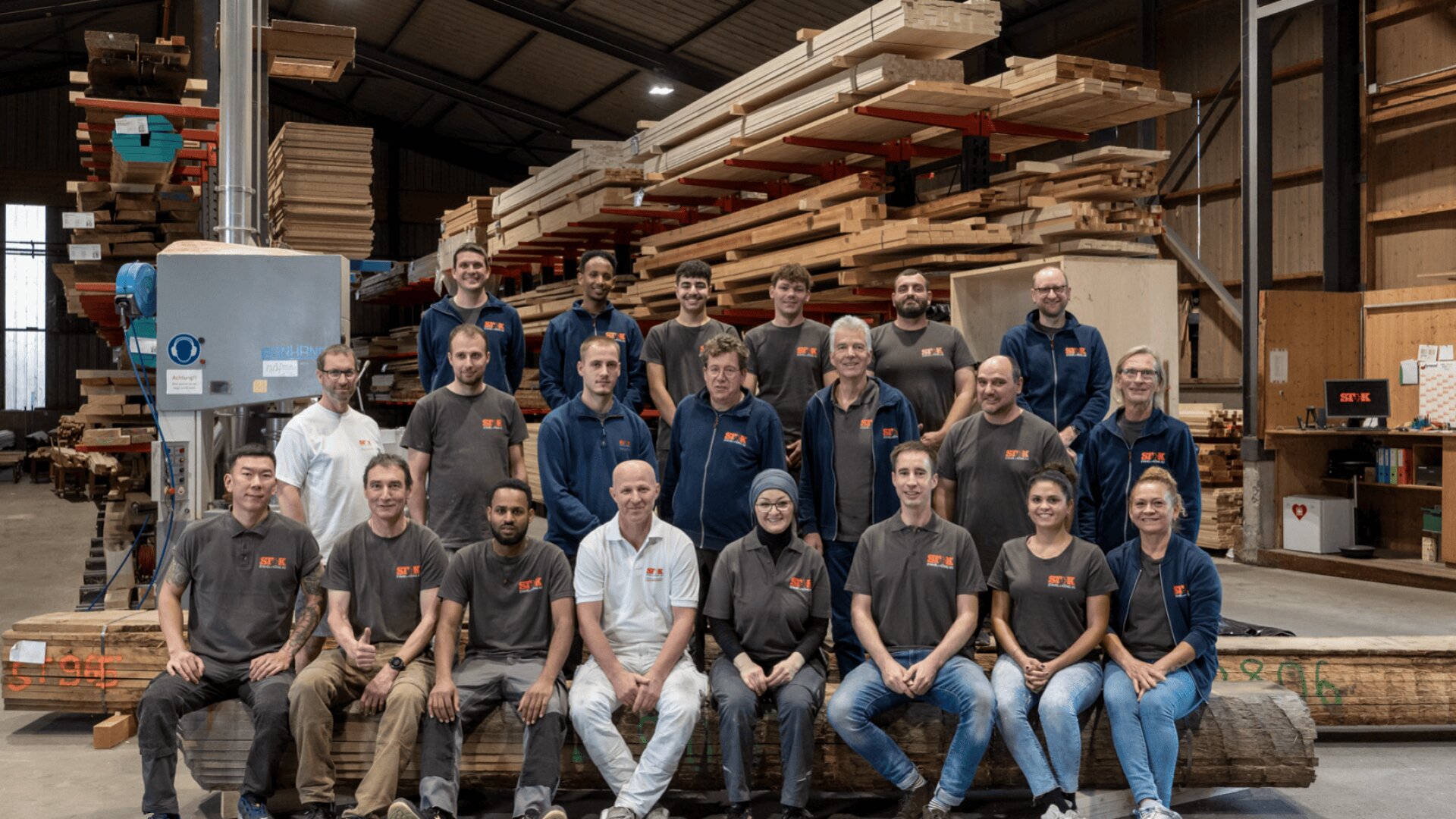 Eine Gruppe von 22 Personen in passenden Arbeitshemden posiert für ein Foto in einer Holzbearbeitungswerkstatt, in der im Hintergrund Holzstapel und Werkzeuge zu sehen sind.