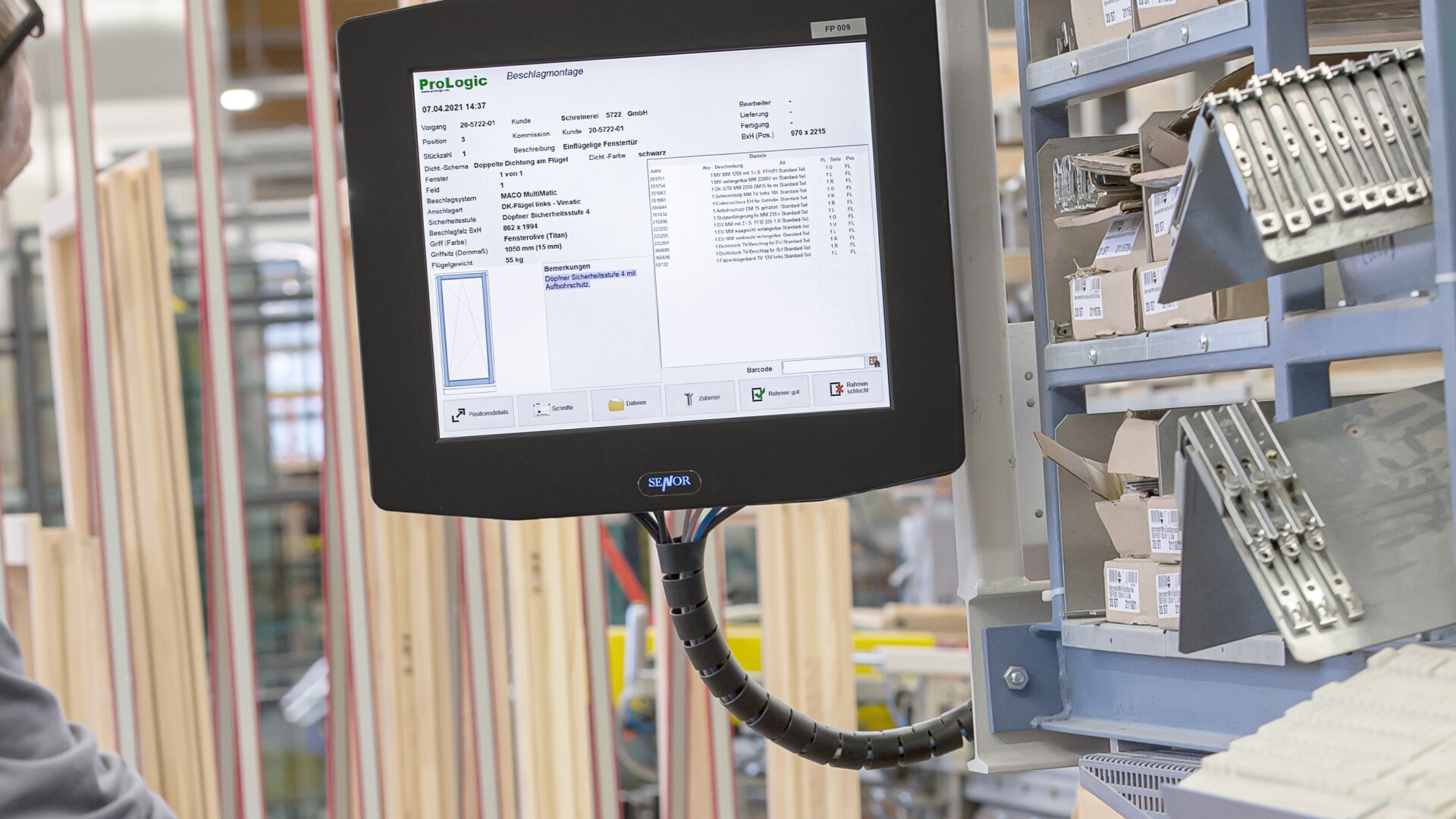 Eine Person mit Maske scannt in einer Werkstatt einen Barcode auf einem Holzrahmen. Im Hintergrund werden auf einem Monitor Bestands- oder Produktionsinformationen sowie Regale mit Materialien angezeigt.