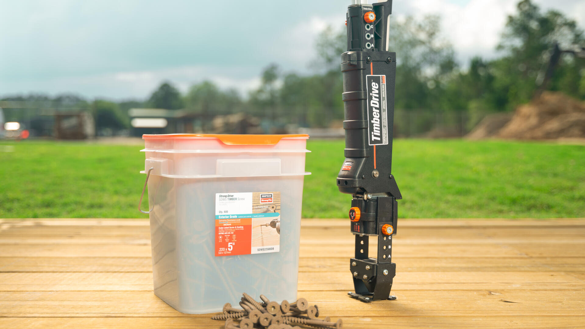Eine Kiste mit Terrassenschrauben und ein Timberdrive-Werkzeug stehen auf einer Holzterrasse im Freien, mit Gras und Bäumen im Hintergrund. Lose Schrauben liegen verstreut vor der Kiste.