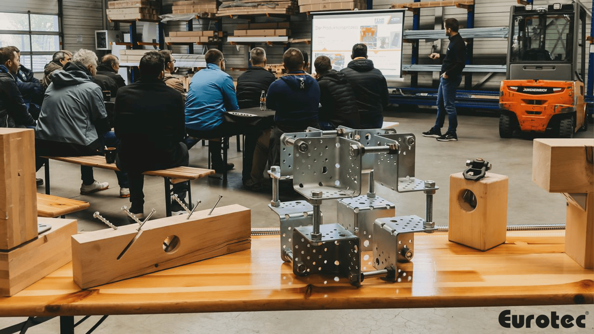 Eine Gruppe von Menschen sitzt auf Bänken in einer Werkstatt und hört einem Vortragenden neben einer Projektionsfläche zu. Im Vordergrund sind Holzbalken und Metallbügel auf einem Tisch ausgestellt. In der Ecke steht das Wort Eurotec.