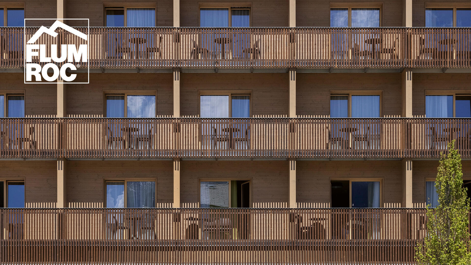 Fassade eines modernen Gebäudes mit Balkonen und Geländern aus Holz, Reihen von grossen Glasfenstern und dem Flumroc-Logo in der oberen linken Ecke. Im Vordergrund unten rechts ist ein Baum zu sehen.
