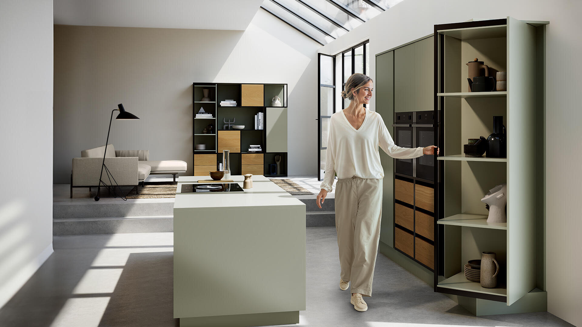 Une femme en vêtements clairs ouvre un haut placard à provisions vert dans une cuisine moderne et minimaliste avec un îlot, des étagères ouvertes et de grandes lucarnes qui laissent entrer la lumière du soleil.