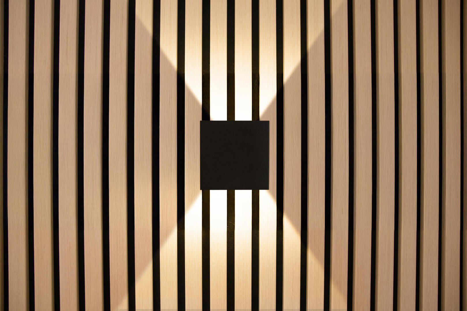 Ein schwarzer, quadratischer Wandleuchter strahlt das Licht in einem X-Muster durch vertikale Holzlatten ab und schafft so ein modernes und geometrisches Lichtdesign.