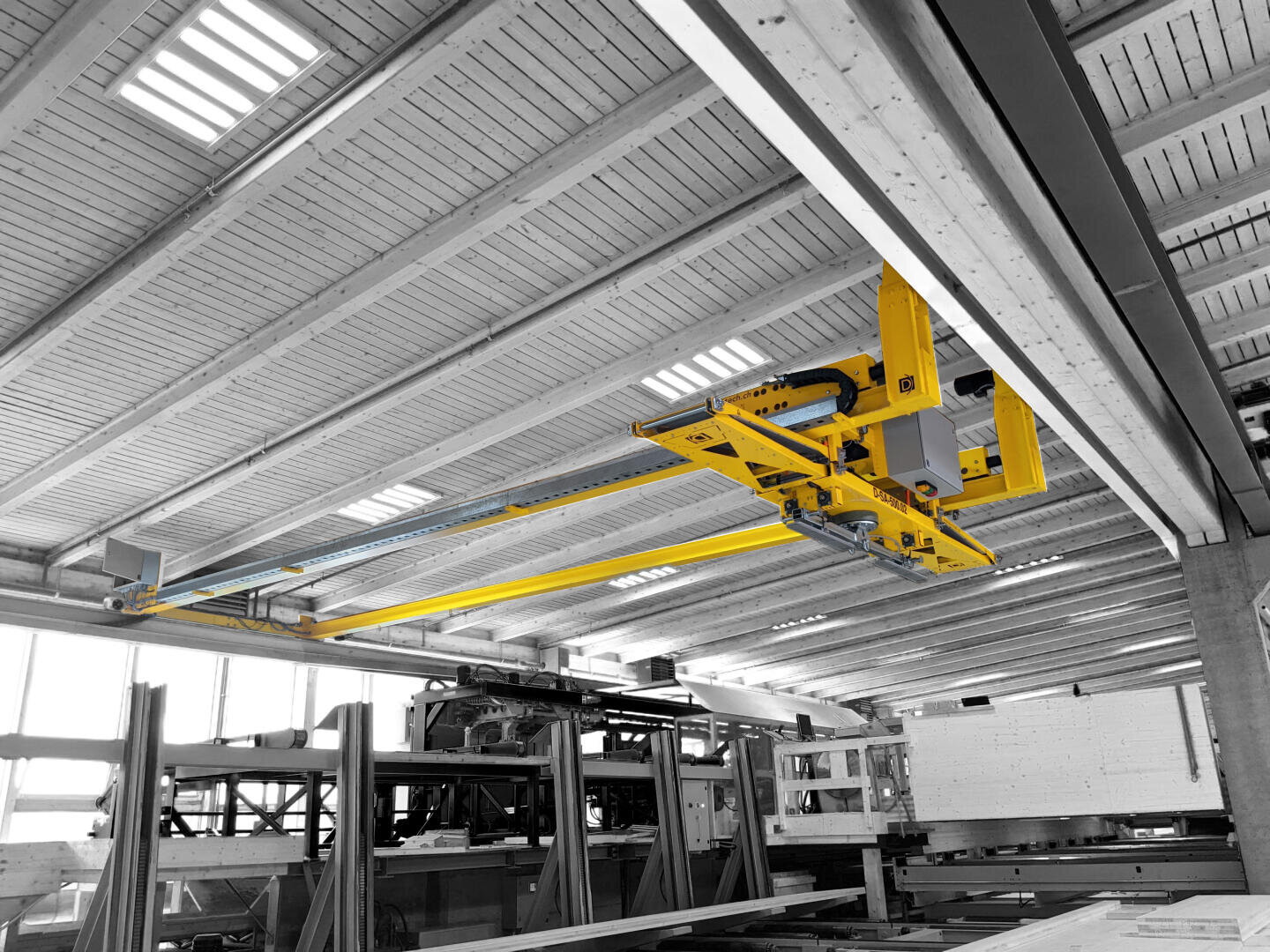 Un système de pont-roulant jaune vif est monté au plafond d'un entrepôt industriel avec des poutres en bois, au milieu de structures en acier et de machines. La majeure partie de l'image est en noir et blanc, mettant en évidence le pont roulant jaune.