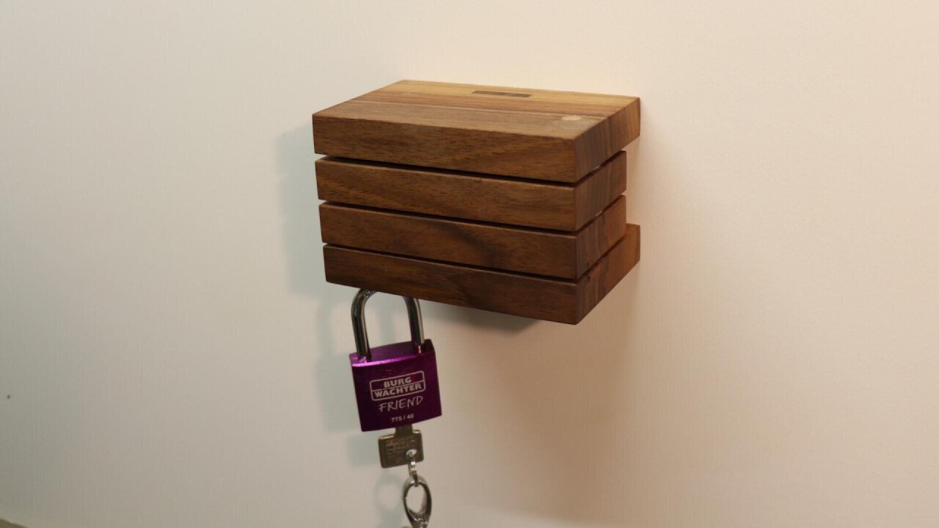 Un porte-clés mural en bois à lamelles horizontales contient un cadenas violet étiqueté FRIEND et un jeu de clés accroché au cadenas. L'arrière-plan est un mur blanc uni.