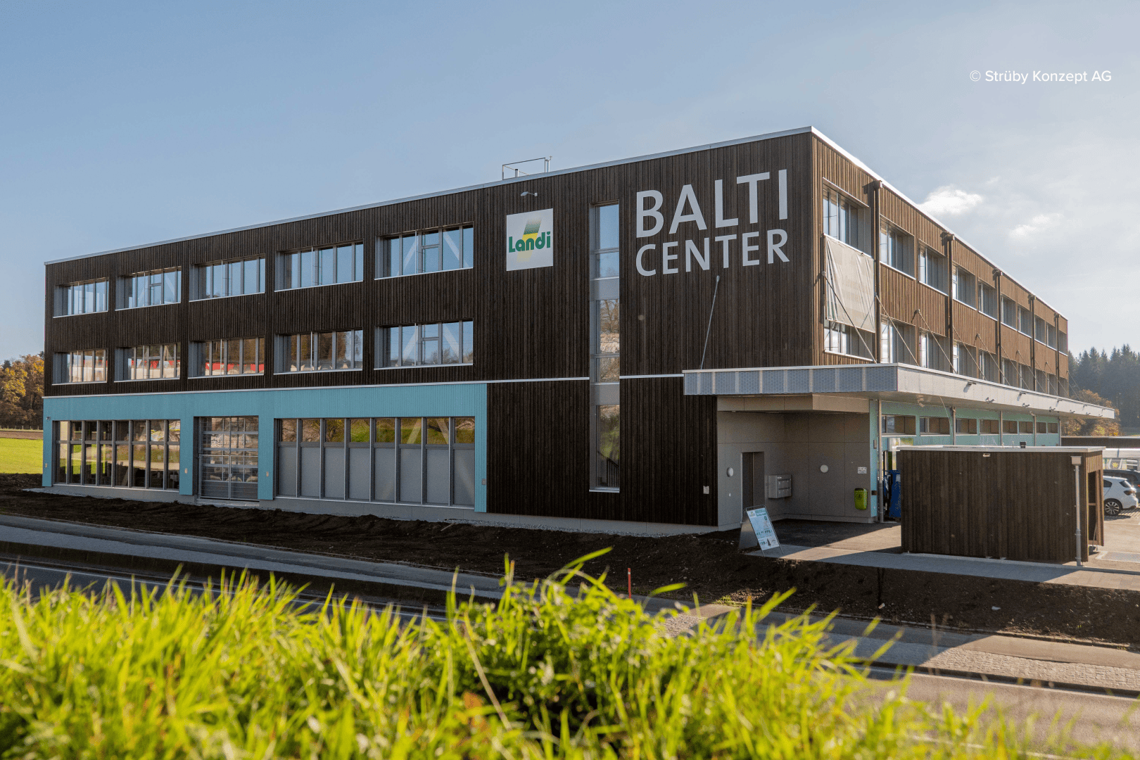 Un bâtiment moderne de trois étages, orné de boiseries et de grandes fenêtres, porte l'inscription « BALTI CENTER ». Une enseigne Landi orne la façade, avec de l'herbe verte et un ciel dégagé au premier plan.