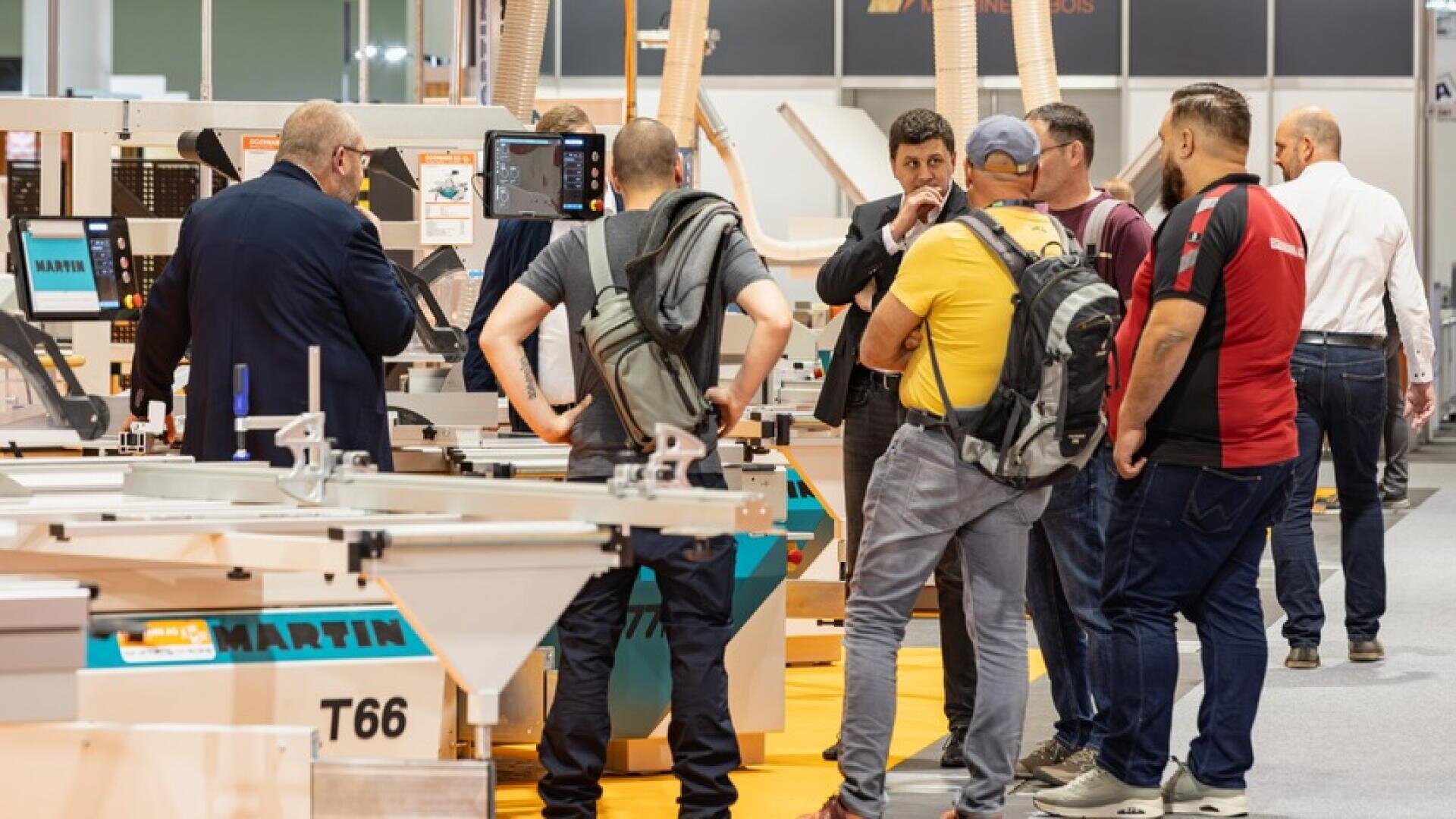 Un groupe d'hommes se tient autour d'une machine à travailler le bois lors d'une exposition et discute. Certains portent des sacs à dos, et divers équipements et écrans sont visibles à l'arrière-plan.