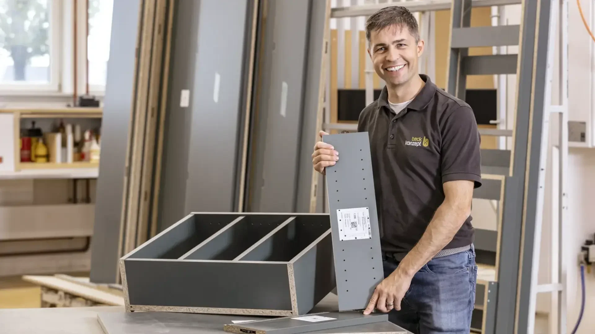 Eine lächelnde Person hält eine graue Platte, während sie in einer Werkstatt ein Möbelstück zusammenbaut, umgeben von verschiedenen Holzteilen und Werkzeugen.